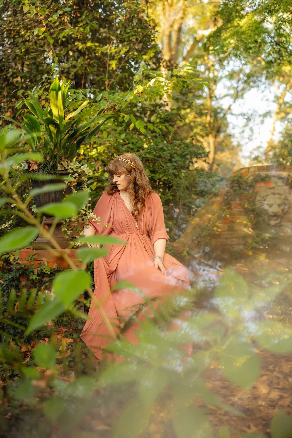 A woman in a pink flowy dress sits among green leafy branches during a photo session in Pike Road, Alabama with m.Clemm photography