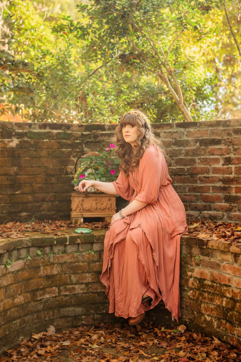 A woman in a pink flowy dress holds a pendulum while sitting on a brick garden wall during a photo session in Pike Road, Alabama with m.Clemm photography