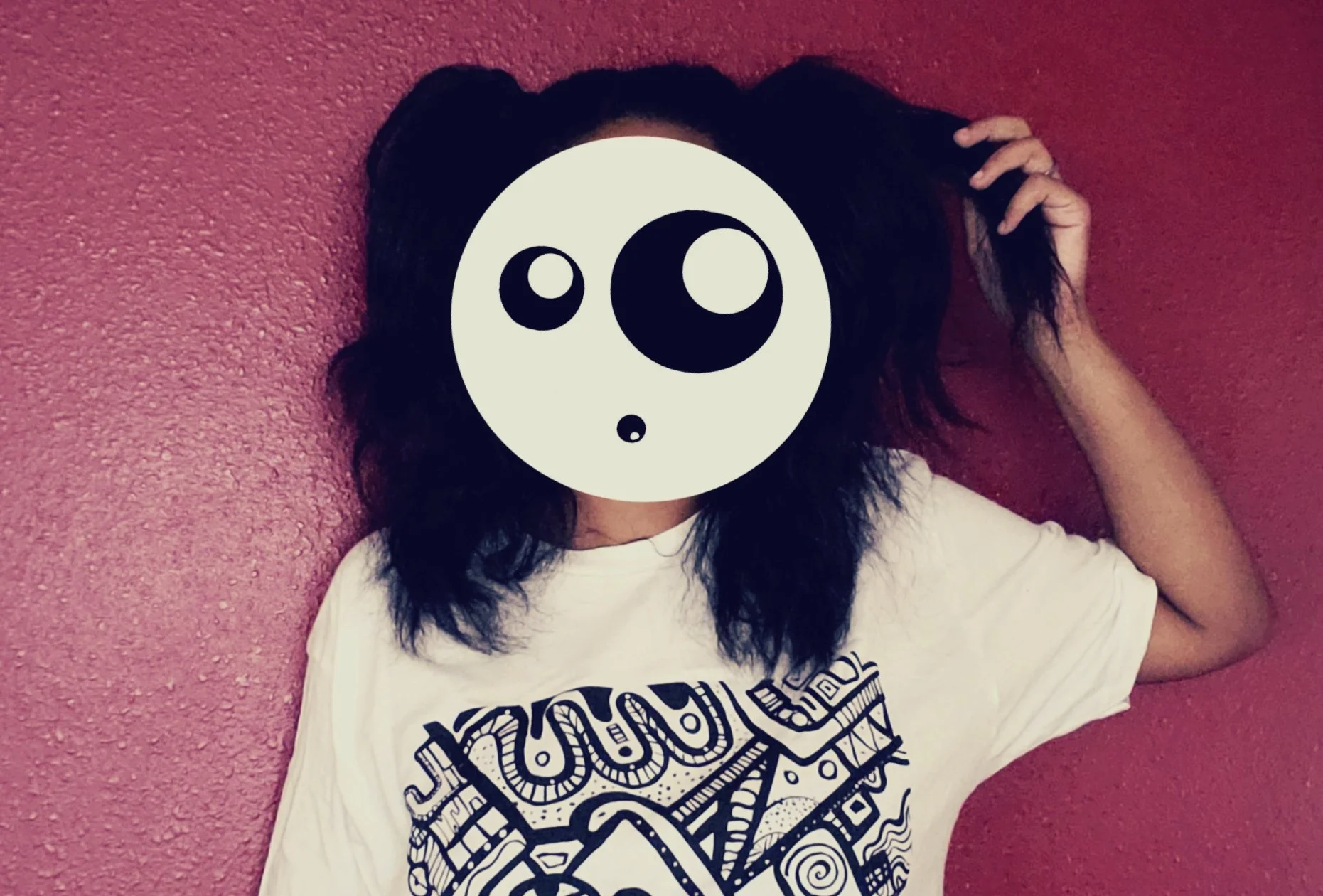 Person with shoulder-length dark hair lying on a pink textured wall, wearing a white graphic t-shirt, with a cartoonish panda face overlay covering the face.