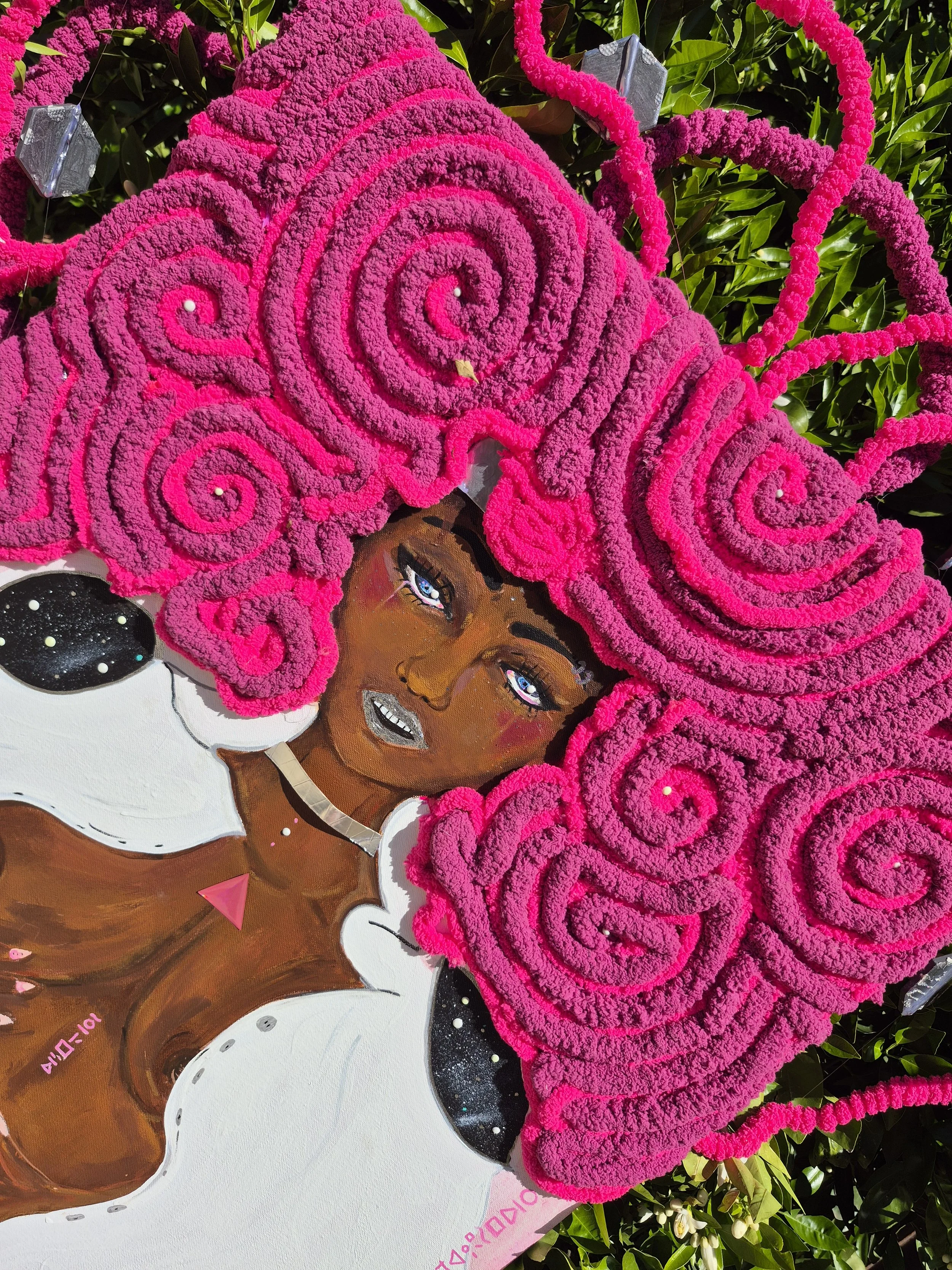 A floral tribute with a painted image of a woman with dark skin, blue eyes, and bright pink curly hair made of pink flowers. The woman is depicted with bold makeup, a necklace, and a confident expression. The tribute is surrounded by greenery.