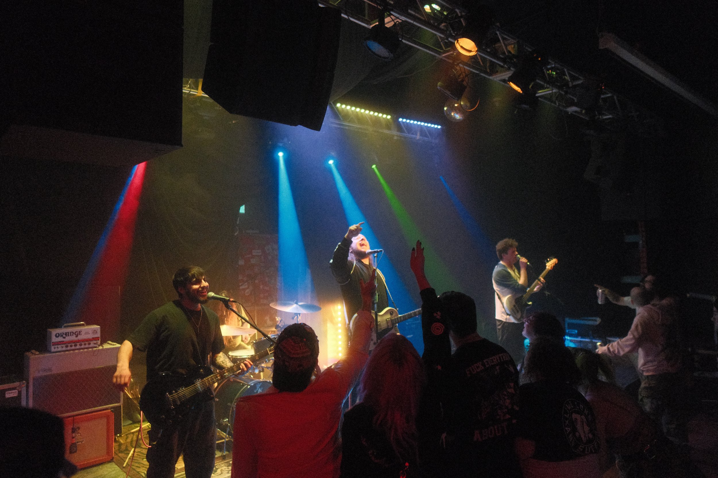 A live musical performance at a concert with musicians playing guitars and singing into microphones on stage, illuminated by colorful stage lights, with a crowd cheering and raising hands.