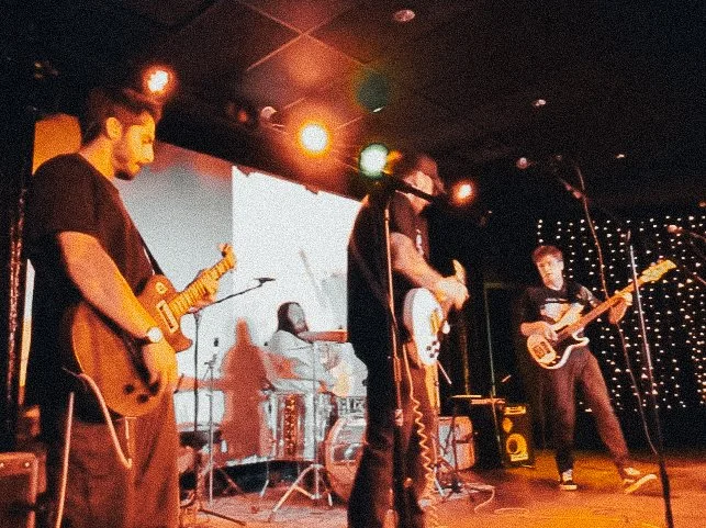 Band of four musicians performing on stage with guitars and drums, set with stage lights and string lights in the background.