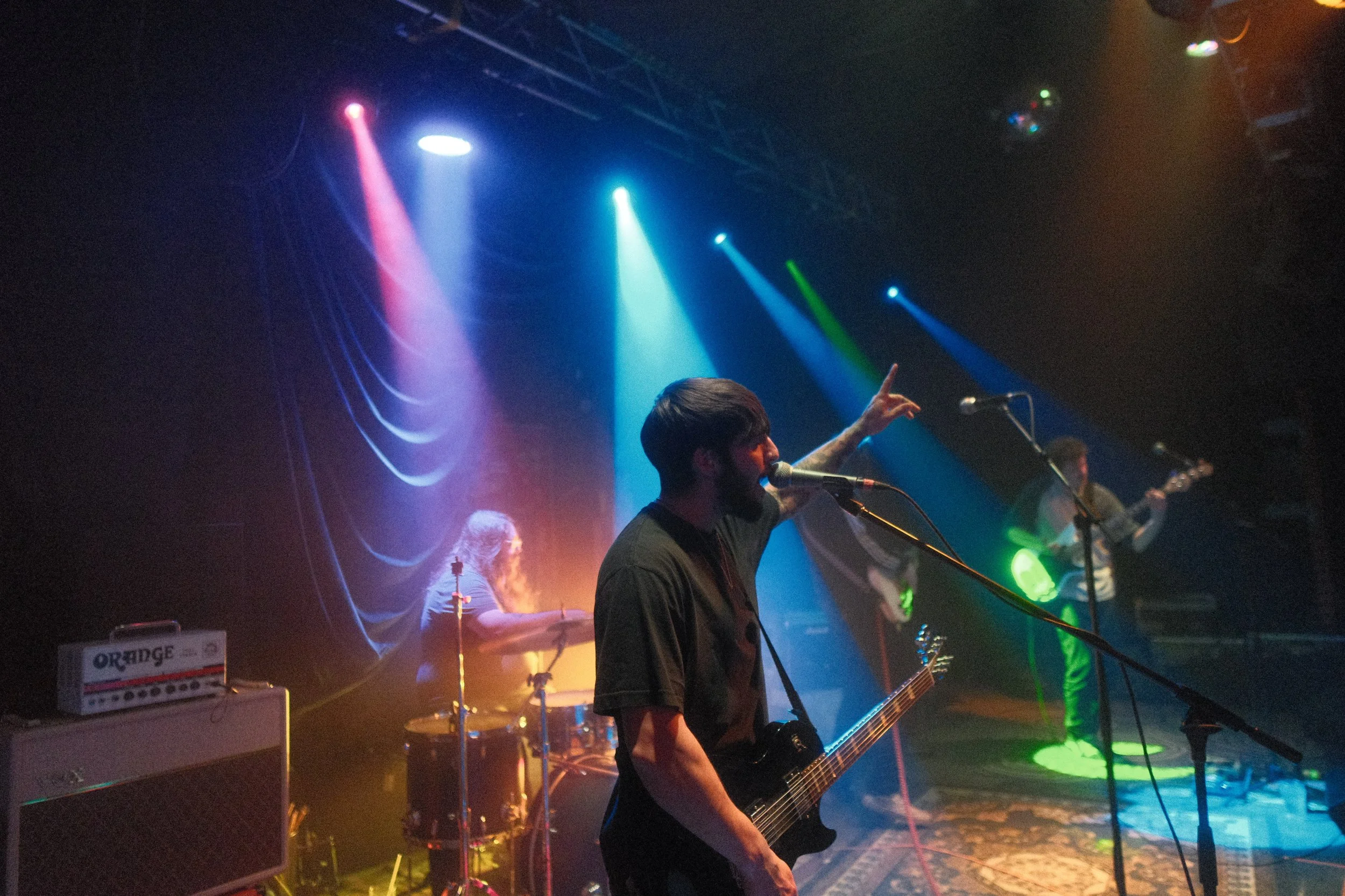 A band performing on stage with colorful stage lighting, including a guitarist singing into a microphone with music equipment around him.