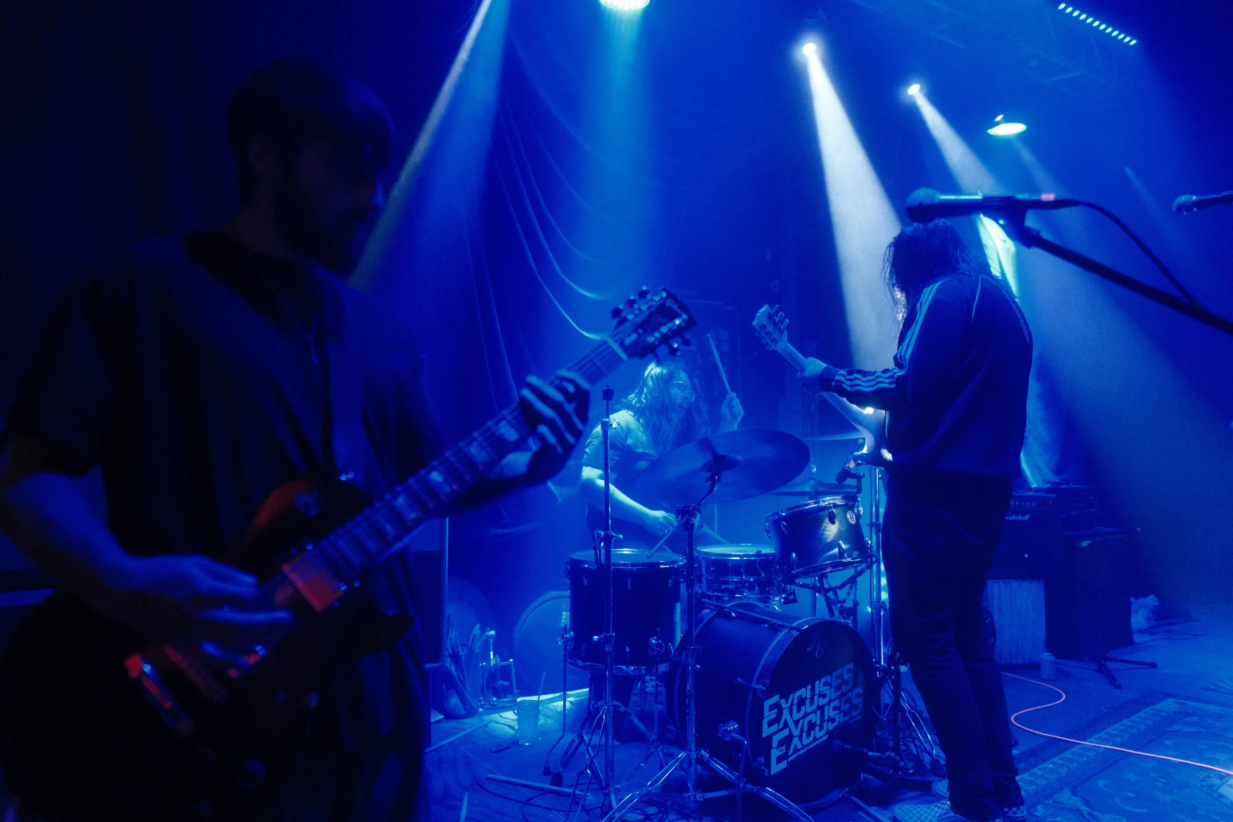 A band performing on stage with blue lighting, featuring musicians playing guitars and drums, with microphones set up in front of them.