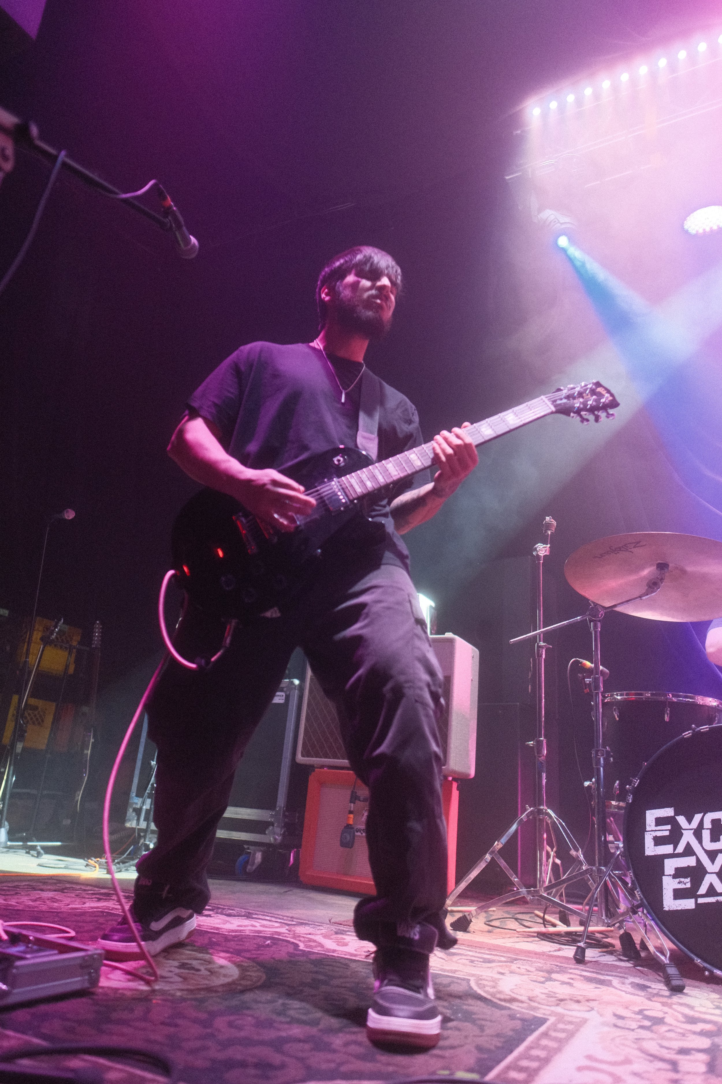 A musician playing an electric guitar on stage during a concert, with purple and pink stage lighting and musical equipment around him.