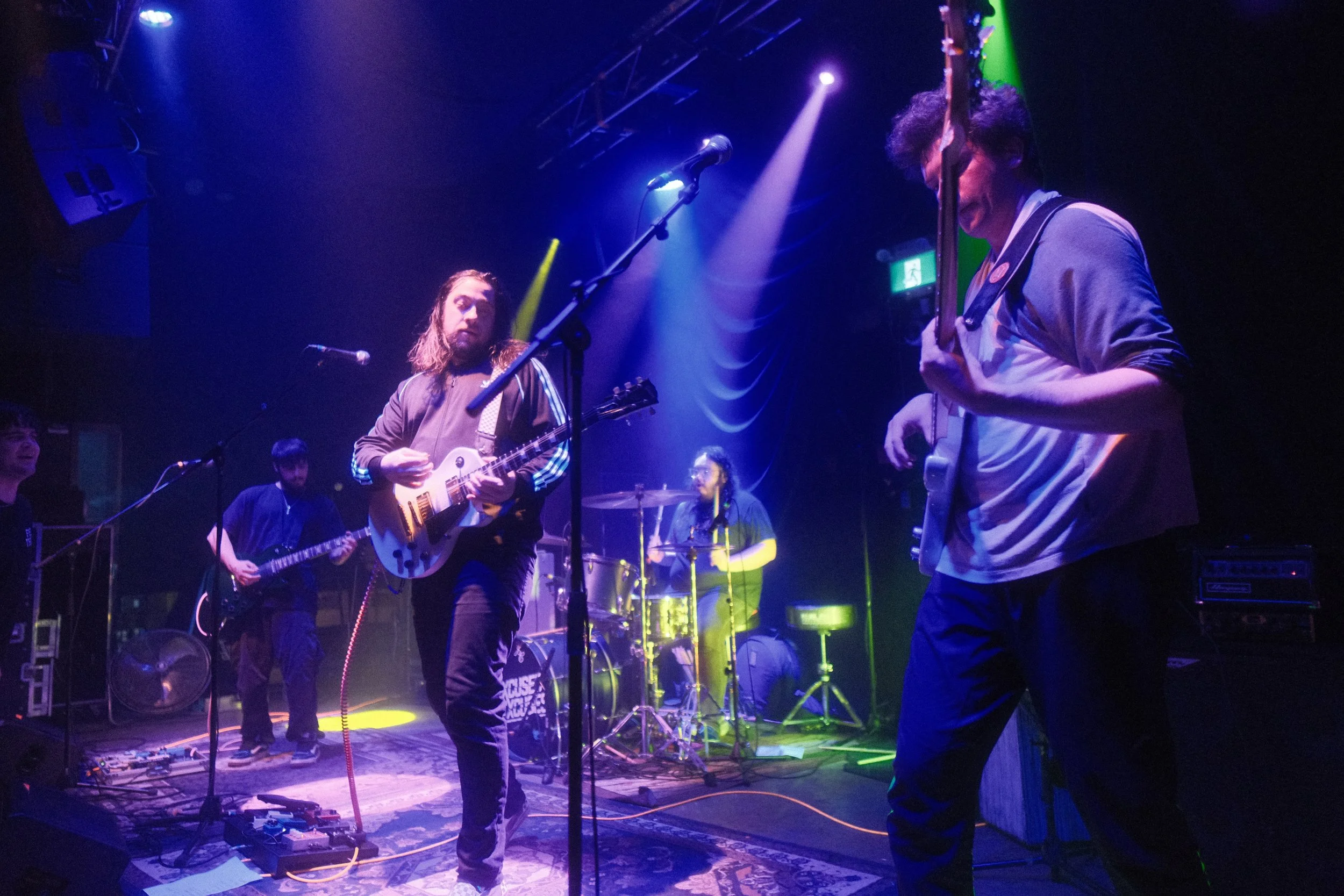 A band performing on stage with colorful lighting, including two guitarists and a drummer, and a few audience members watching.