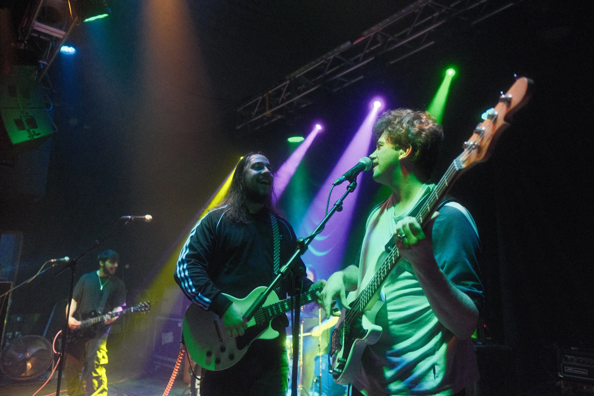 Two musicians playing guitars and singing on stage with colorful stage lights overhead, background performers, and stage equipment.