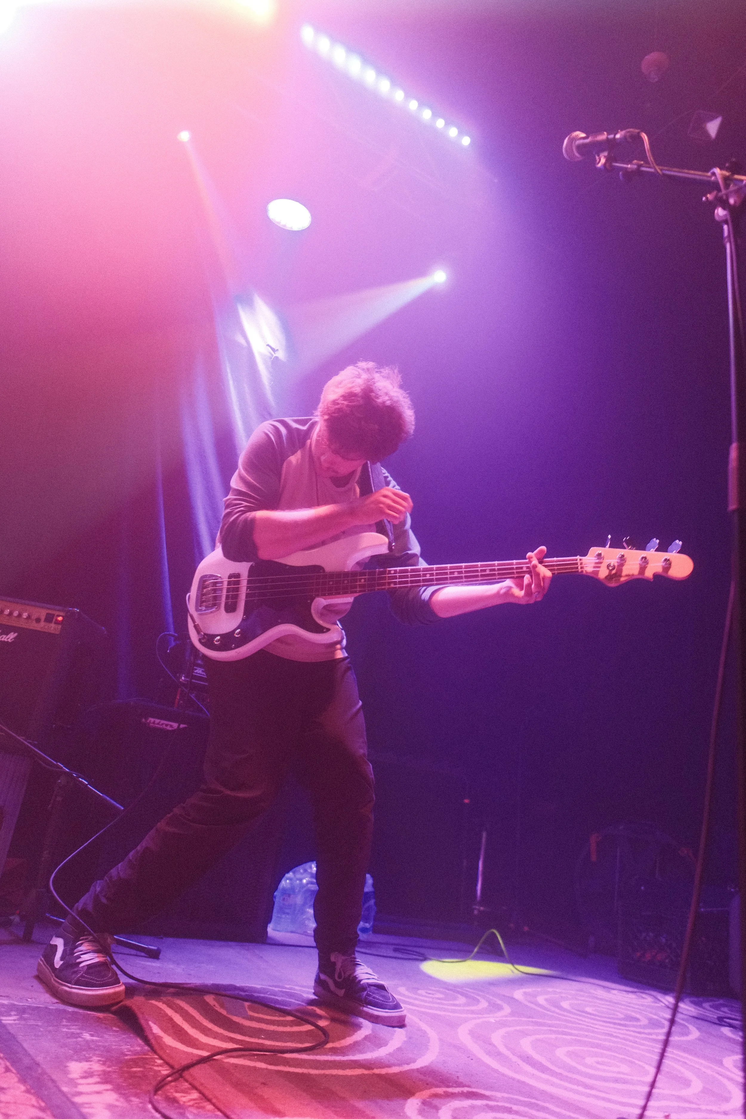 Musician playing bass guitar on stage with colorful stage lighting, wearing casual clothing and sneakers.