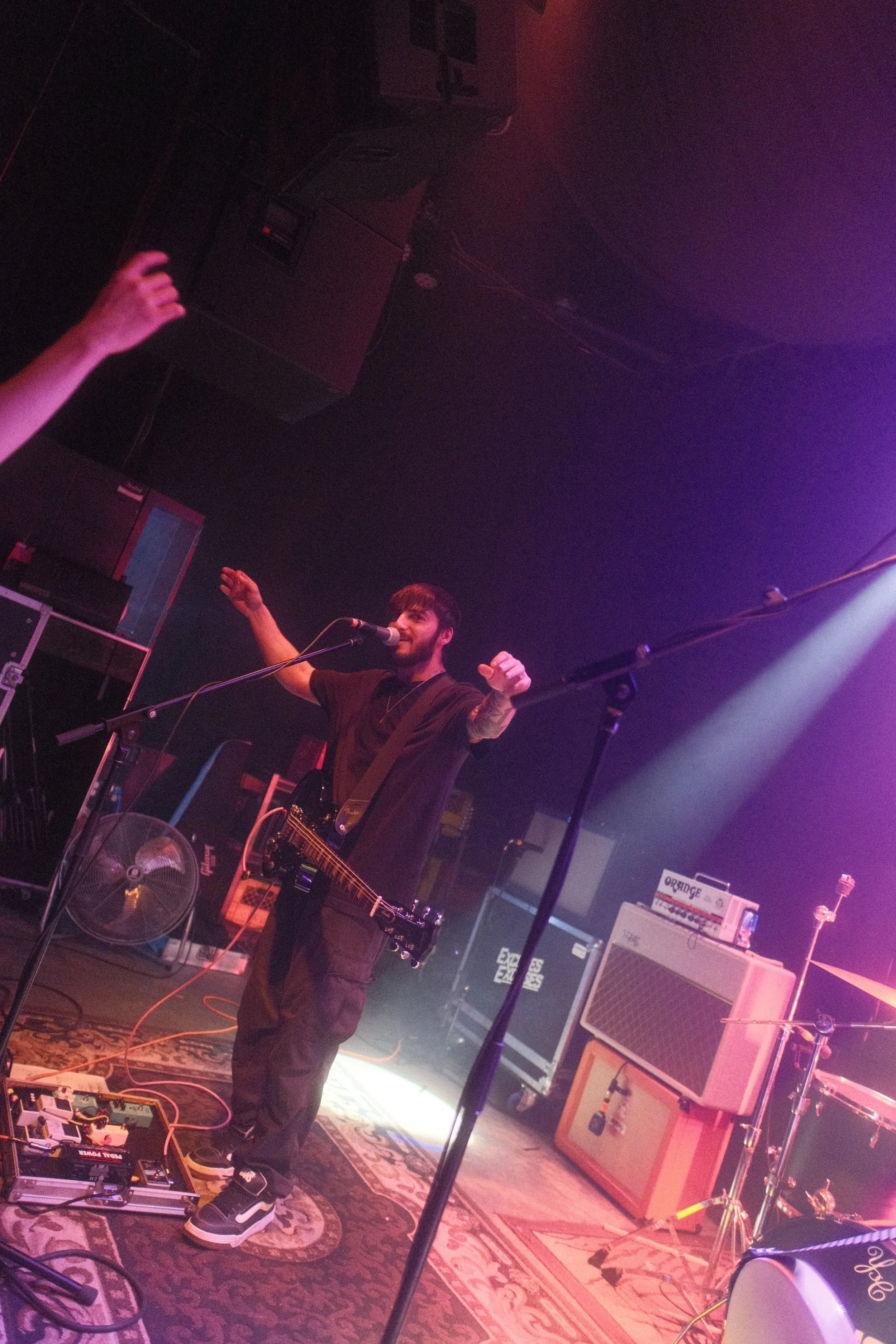A musician performing on stage with a guitar and microphone, raised hands in the air, surrounded by amplifiers and musical equipment, in a dimly lit venue with purple and pink lights.
