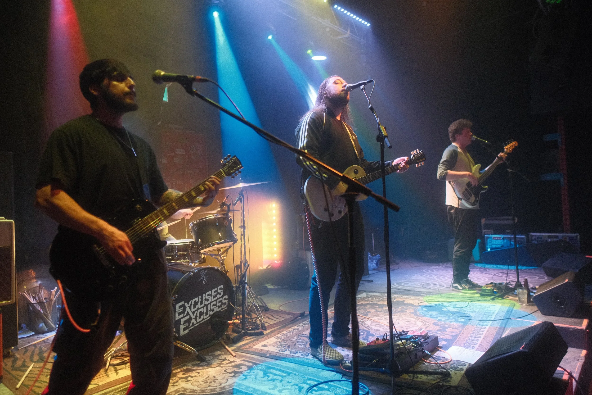 A band performs on stage with colorful stage lights, featuring three guitarists and a drum set, at a live music venue.