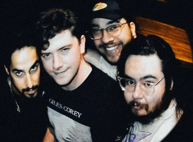 Four men smiling for a selfie, three with glasses, dark hair, and facial hair. They appear to be in a dimly lit indoor setting.