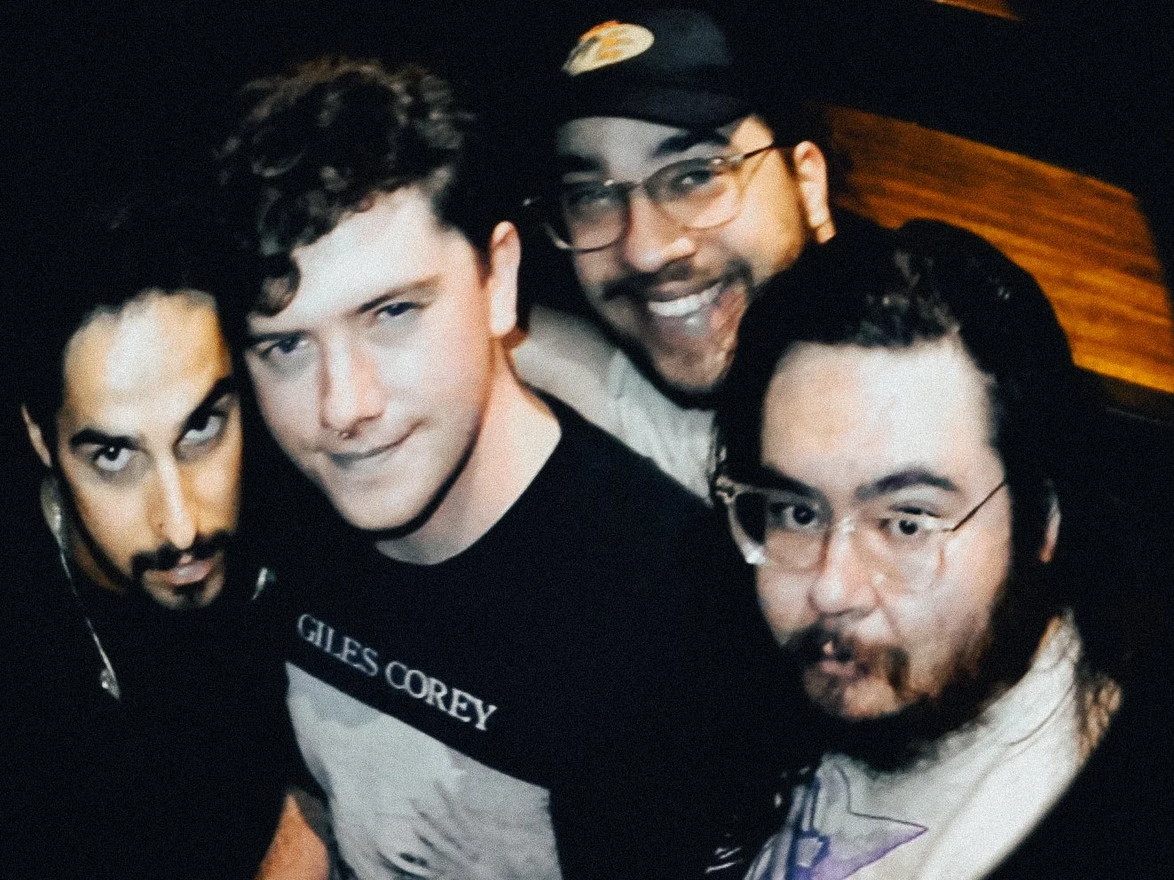 Four men with dark hair and facial hair, posing closely together for a selfie, with a wooden wall in the background.