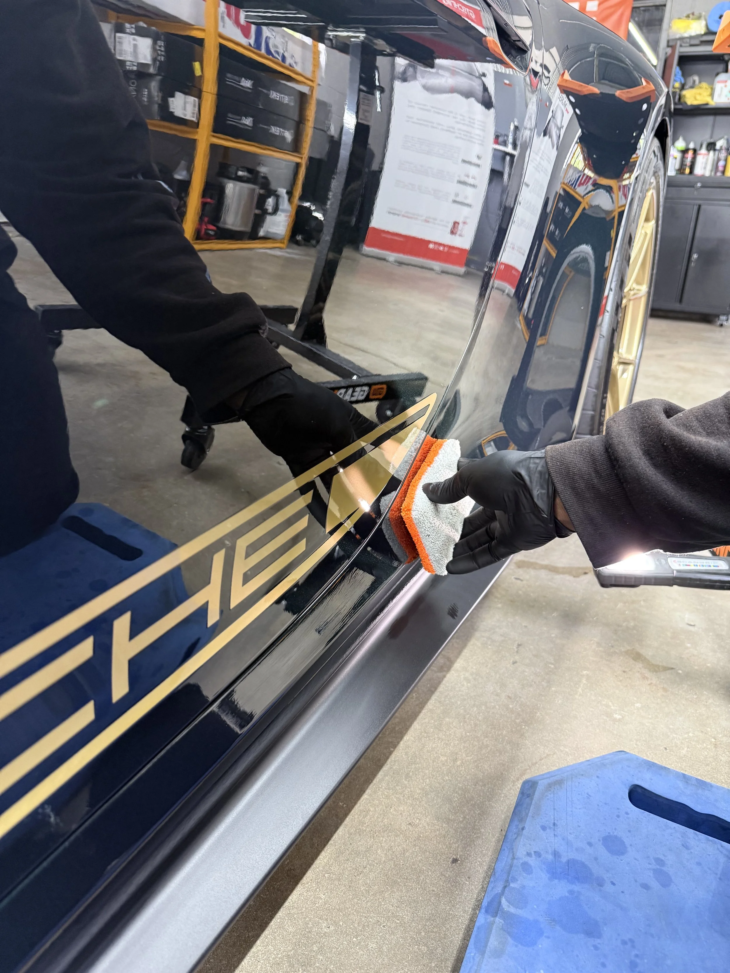 Professional detailer polishing a black car with a towel in a garage or workshop.