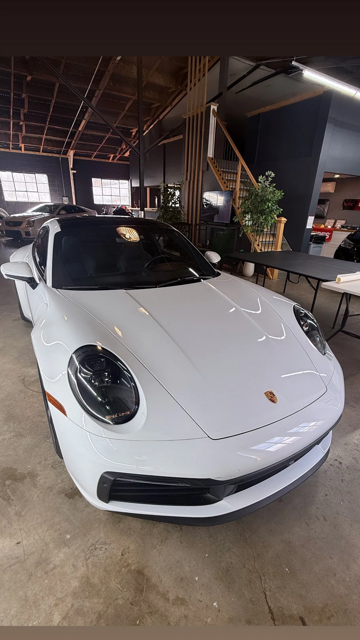 A white Porsche sports car displaying Paint Protection Film inside a showroom with other cars visible in the background and a staircase leading to an upper level.