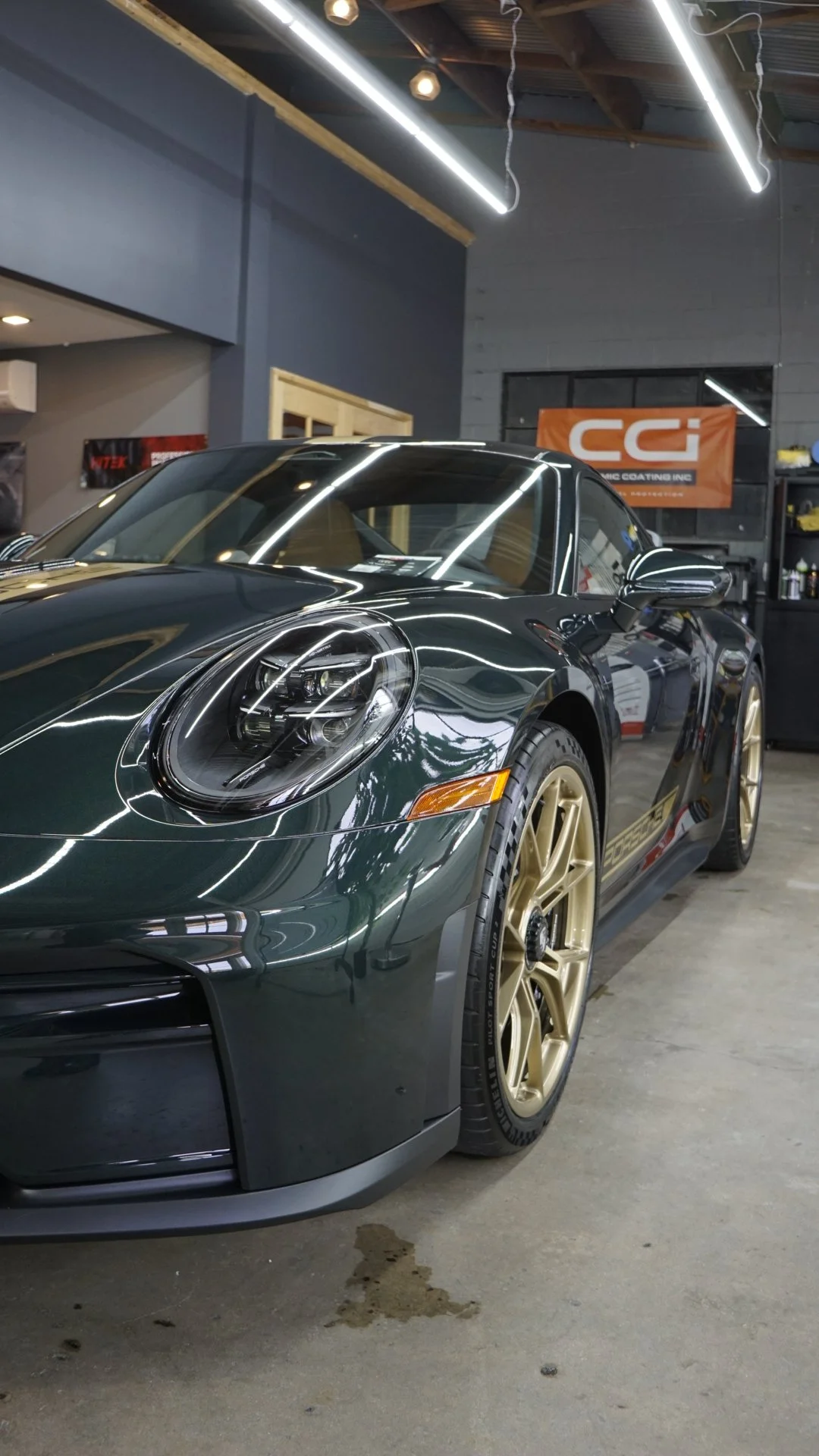 A dark green Porsche sports car with gold wheels parked inside a garage with gray walls and exposed ceiling beams, reflected lights on the car's surface, and a sign that reads Carrera.