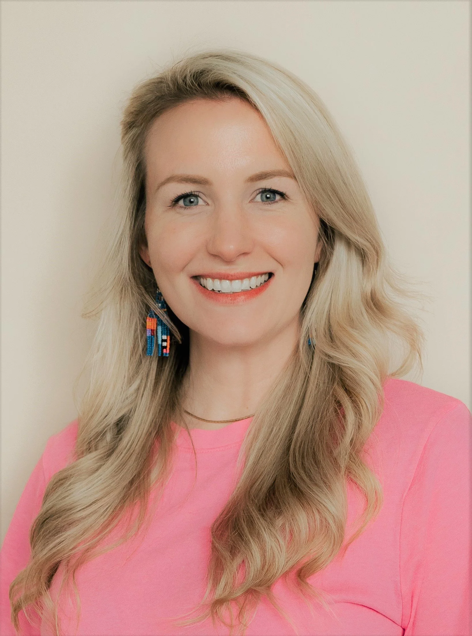 A smiling woman with long blonde hair, wearing a pink top and beaded earrings, against a plain wall background.