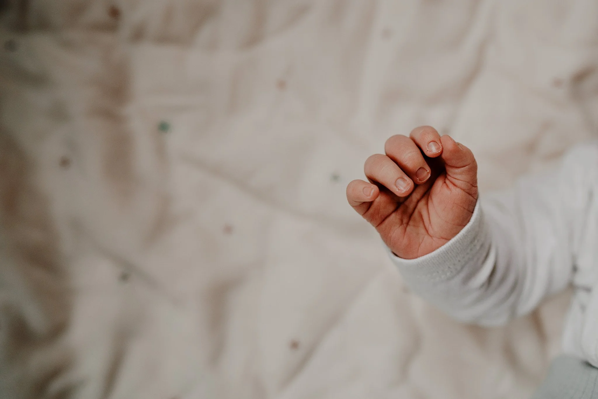 Close-up of a small child's hand with fingers slightly curled, wearing a white long-sleeve garment, against a soft, neutral background.