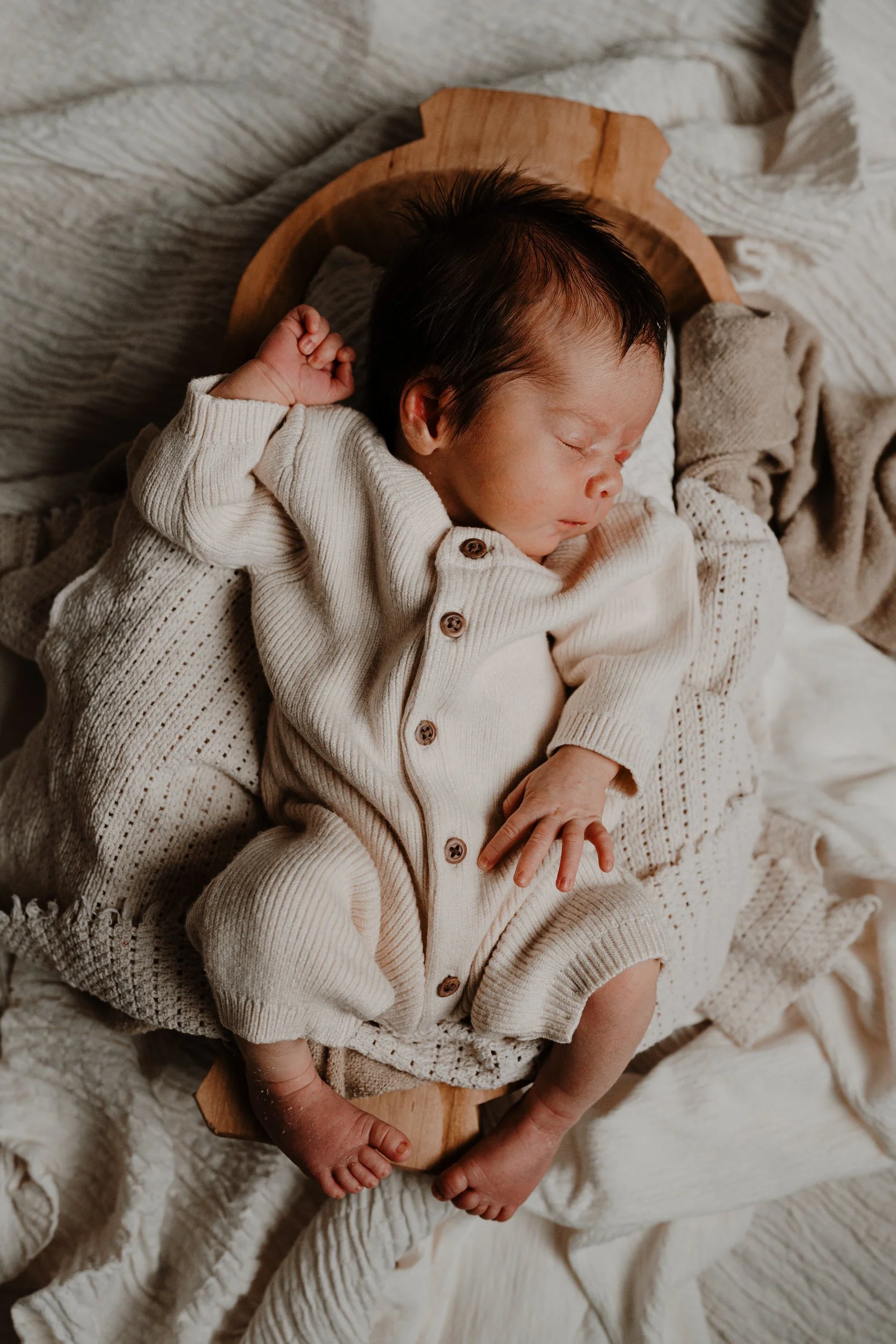 A sleeping baby with black hair, dressed in a beige knitted onesie, lying on a wooden tray with blankets around, on a bed with cream-colored linens.