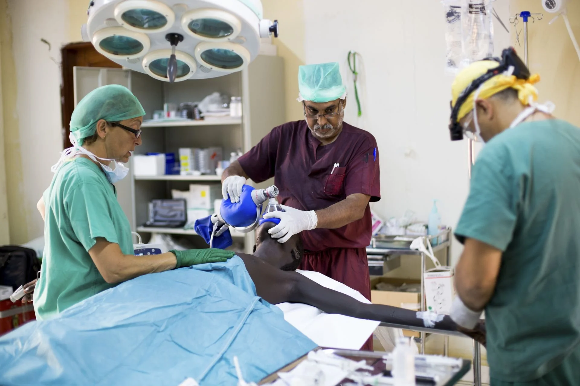An anaesthesia provider holds a mask over a patient’s face and squeezes a blue manual ventilation bag on an operating table, while two members of the surgical team stand on either side.