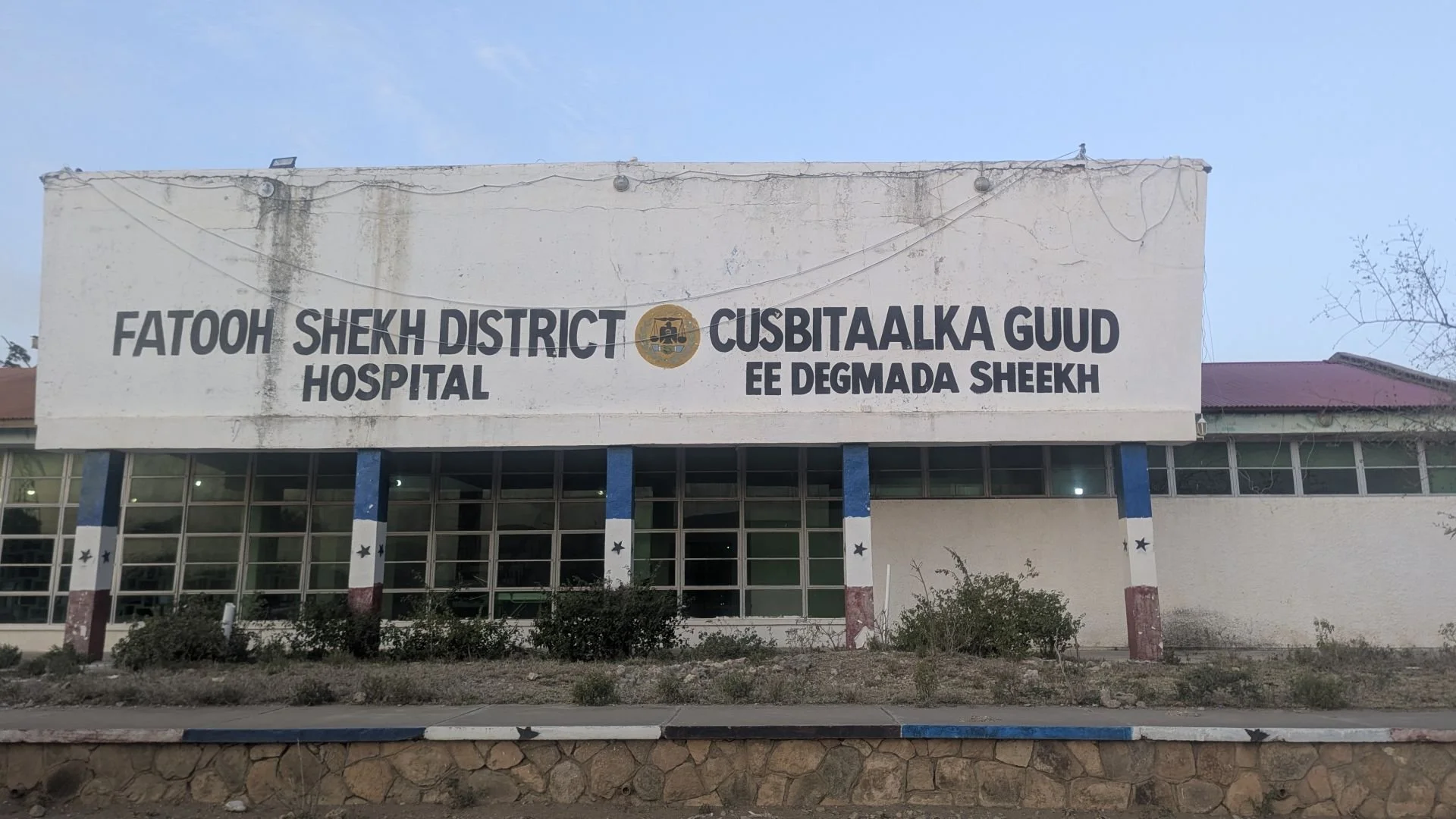 Front of Sheikh District Hospital in Somaliland, its sign reading Fatooh Shekh District Hospital in English and Cusbitaalka Guud ee Degmada Sheekh in Somali