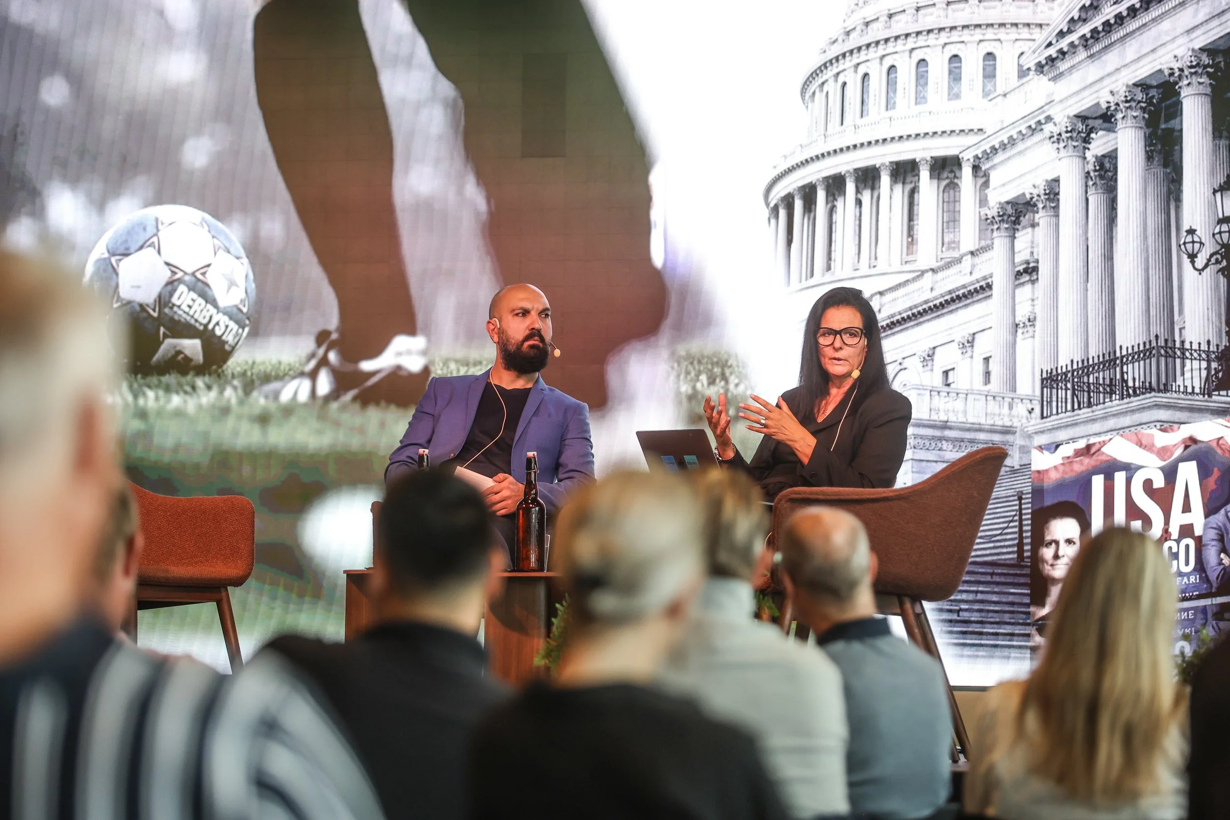 Två personer talar på en paneldebatt, med ett publikhav framför sig. Bakgrunden visar en modern byggnad och ett fotbollsspel.
