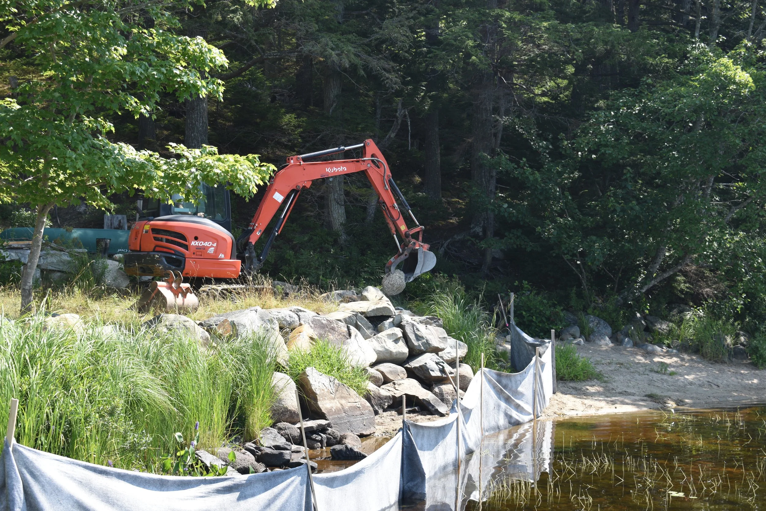 A shoreline with silt fencing, rock wall, and an orange excavator removing rock.
