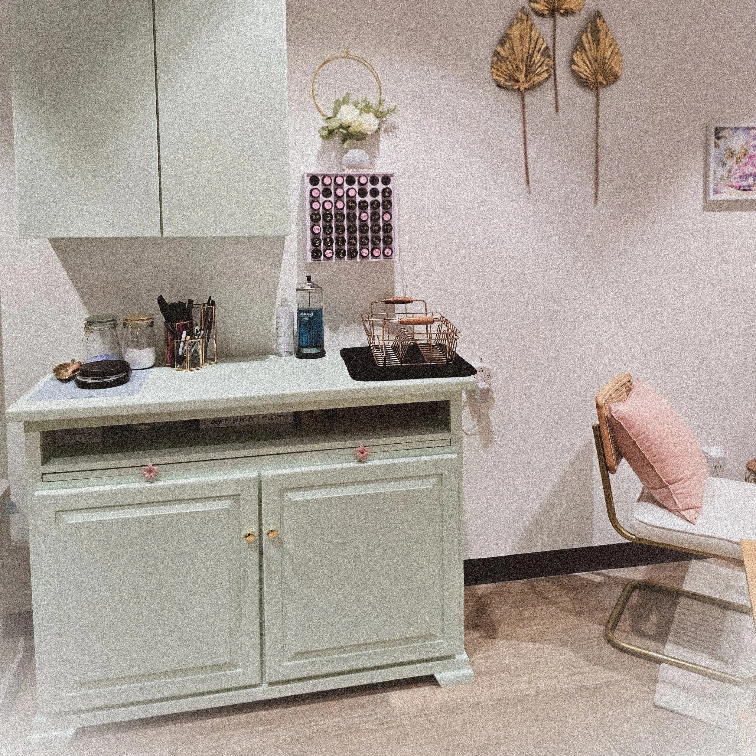 A cozy corner featuring a vintage white cabinet with pink floral handles, a small chair with a pink pillow, and wall decor including golden leaf ornaments and a framed picture. The cabinet top displays a small vase with white flowers, a wine rack, and small jars.