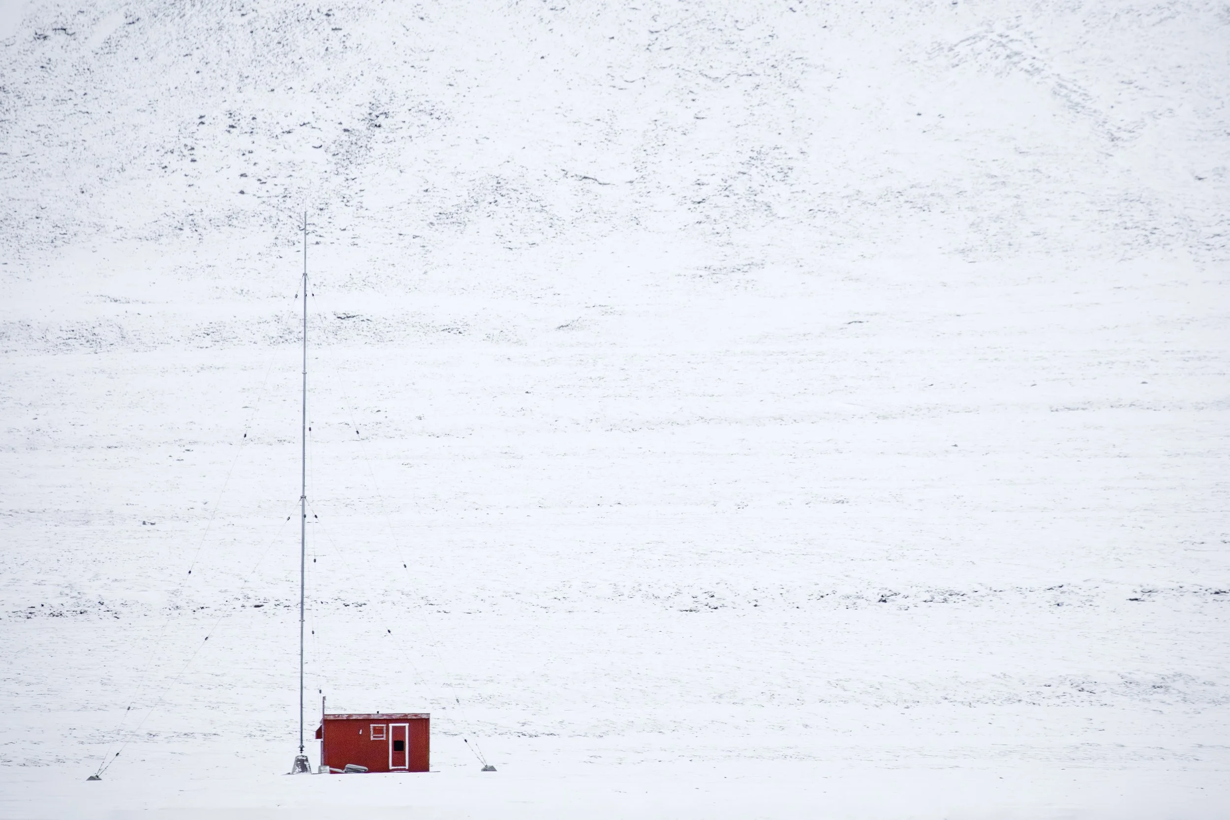 A small red building or hut with a door and window, situated on a snowy landscape, with a tall, thin antenna extending upward from the building.