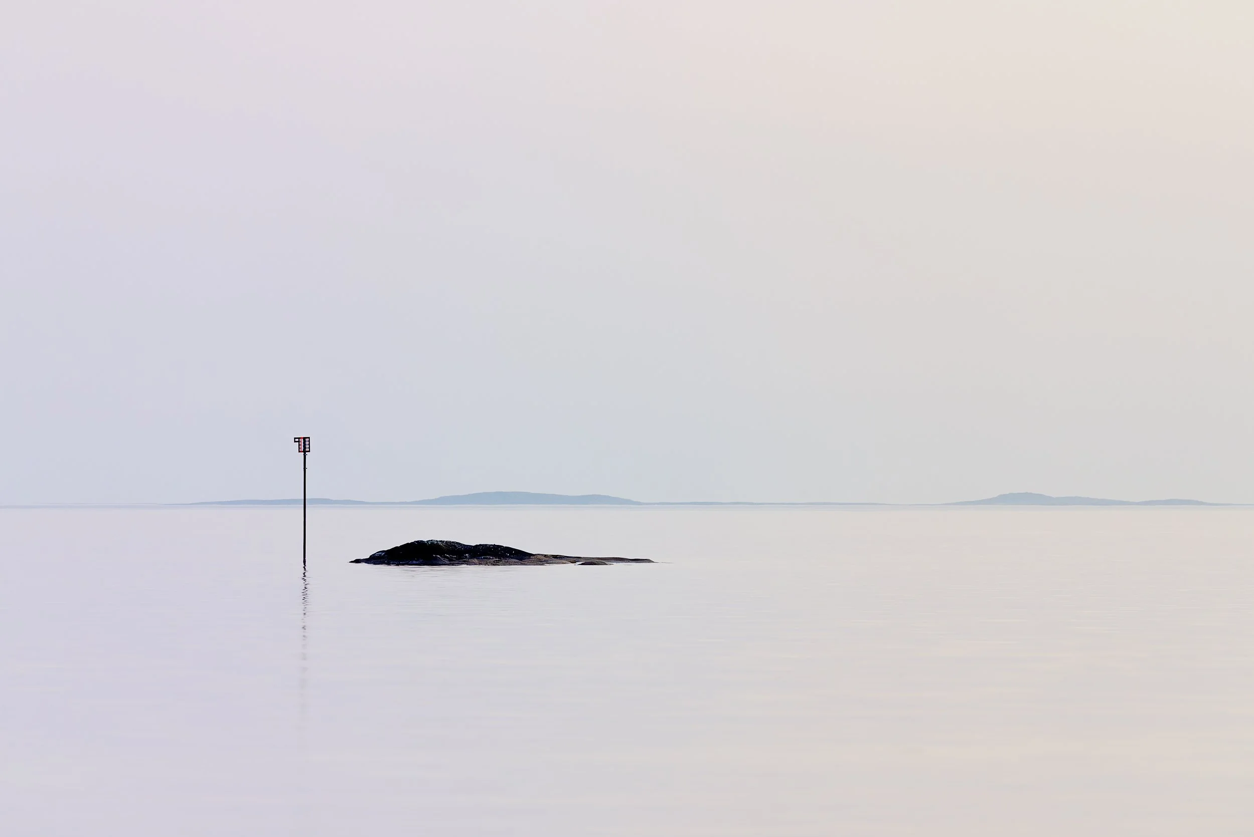Calm sea with a small island, a tall navigation marker, and distant landforms in the background under a cloudy sky.