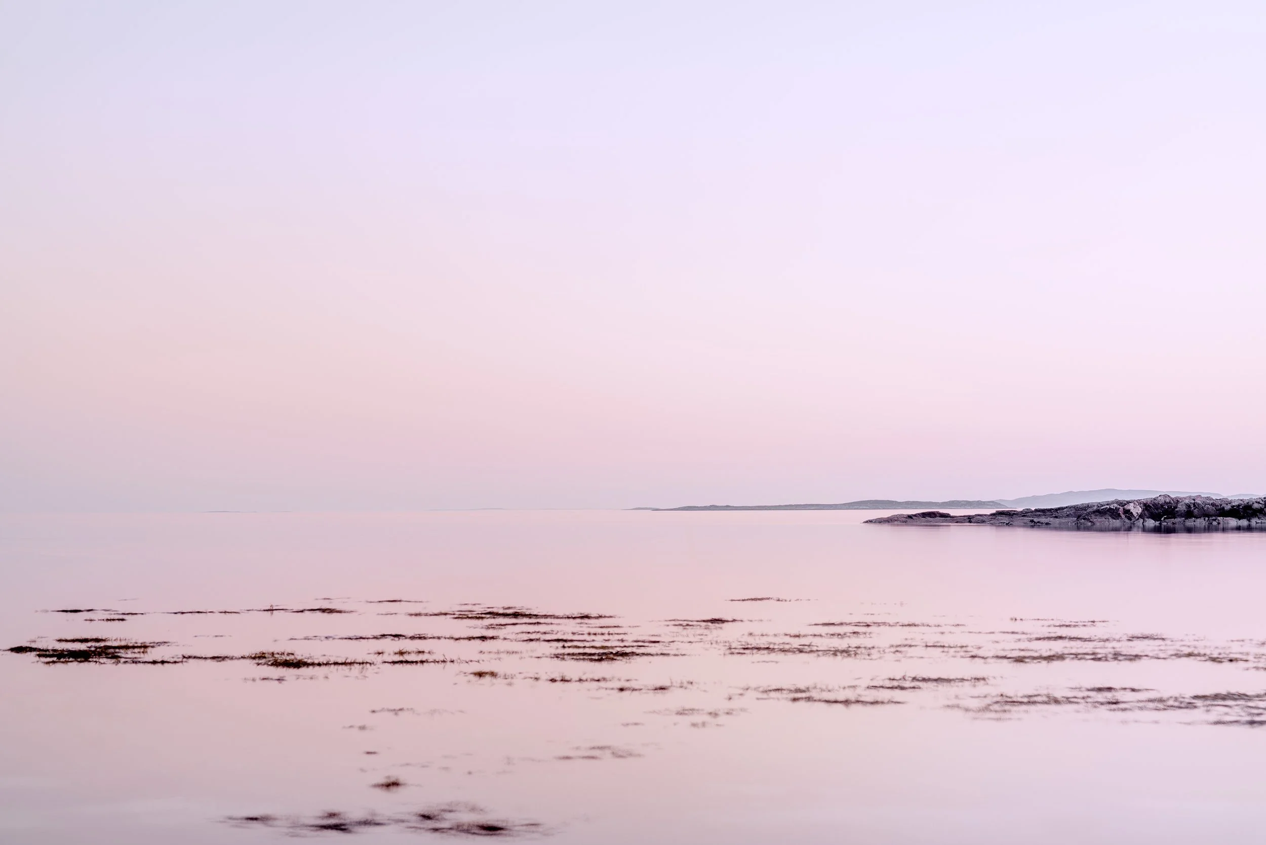 Calm pink and purple pastel sunset over a tranquil body of water with rocks on the right side.