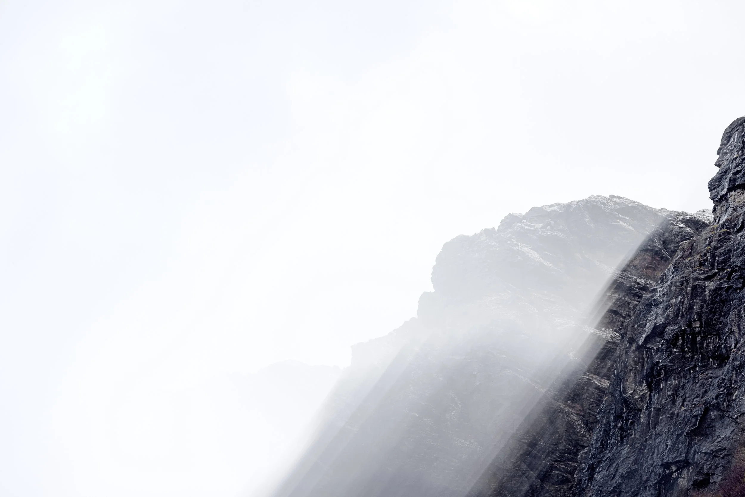 Light rays cascading down dark rocky mountain slopes with mist and a gray, overcast sky