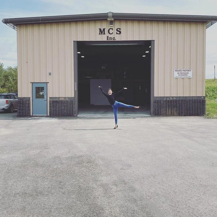 A woman dressed in black with blue jeans and high heels is performing a ballet pose, balancing on one leg with her arms raised and the other leg extended behind her, in front of a large warehouse with the sign 'MCS Inc.' on the front.