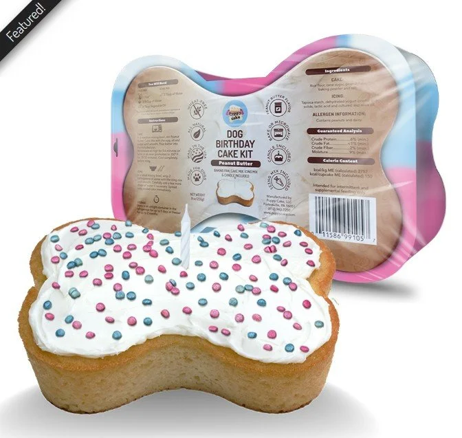 Dog-shaped birthday cake with white frosting and pink and blue sprinkles, standing in front of its packaging labeled 'Dog Birthday Cake Kit' and 'Peanut Butter'.