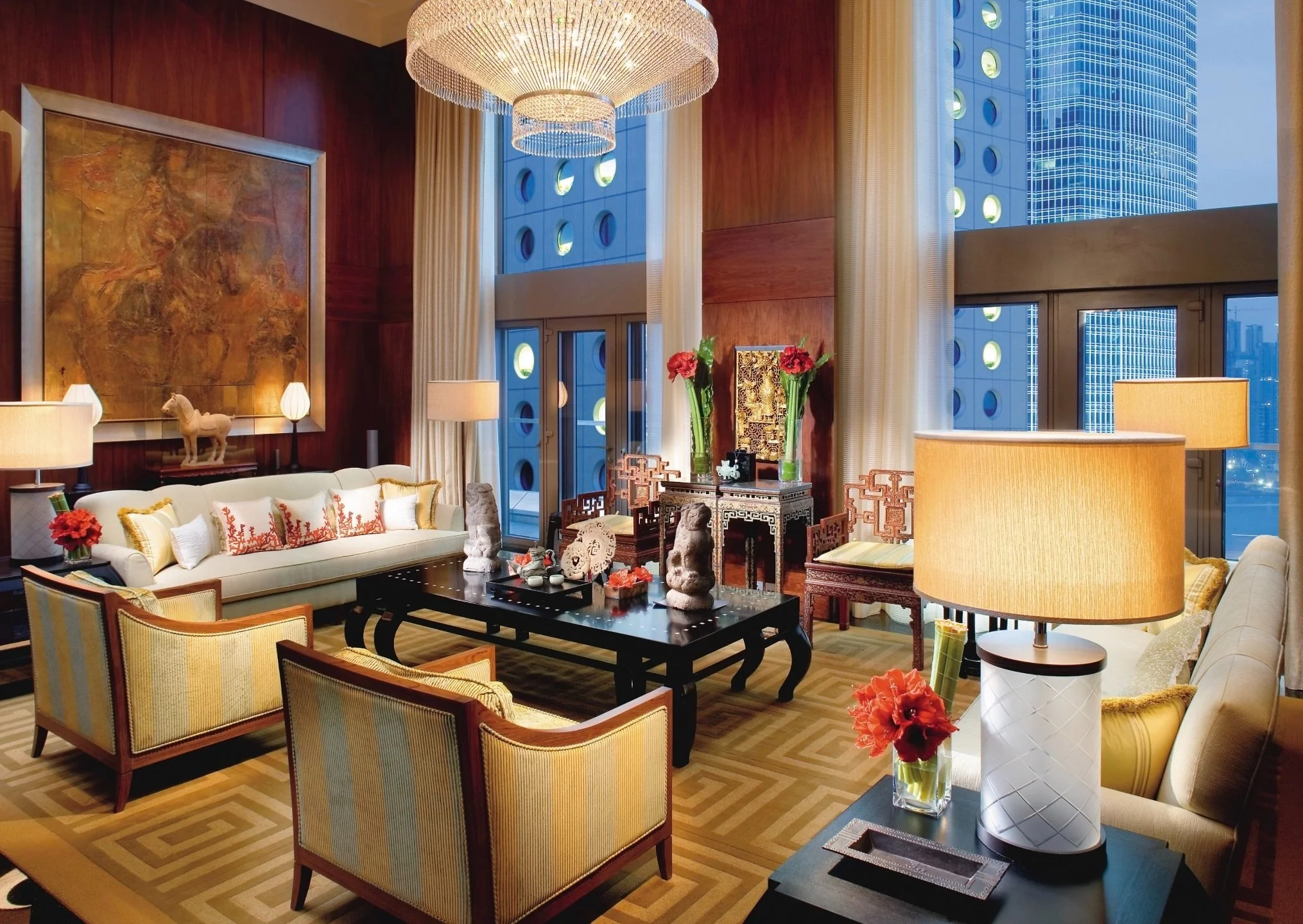 Elegant living room with cream sofas, yellow armchairs, and a black coffee table, decorated with vases of red flowers, sculptures, and art pieces, with large windows and city skyline outside.