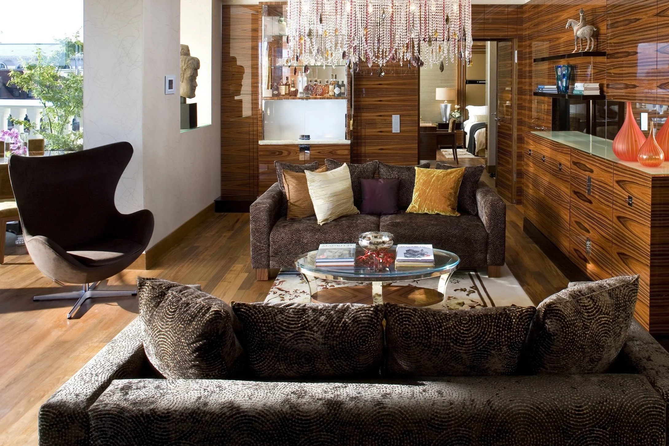 Living room with dark patterned sofas, glass coffee table, black modern armchair, wooden wall panels, chandelier, shelves with decorative items, and a view into a bedroom.