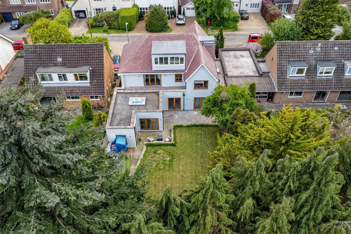 An aerial view of a modern house with a gray roof and white walls, surrounded by trees and neighboring houses, in a suburban neighborhood.