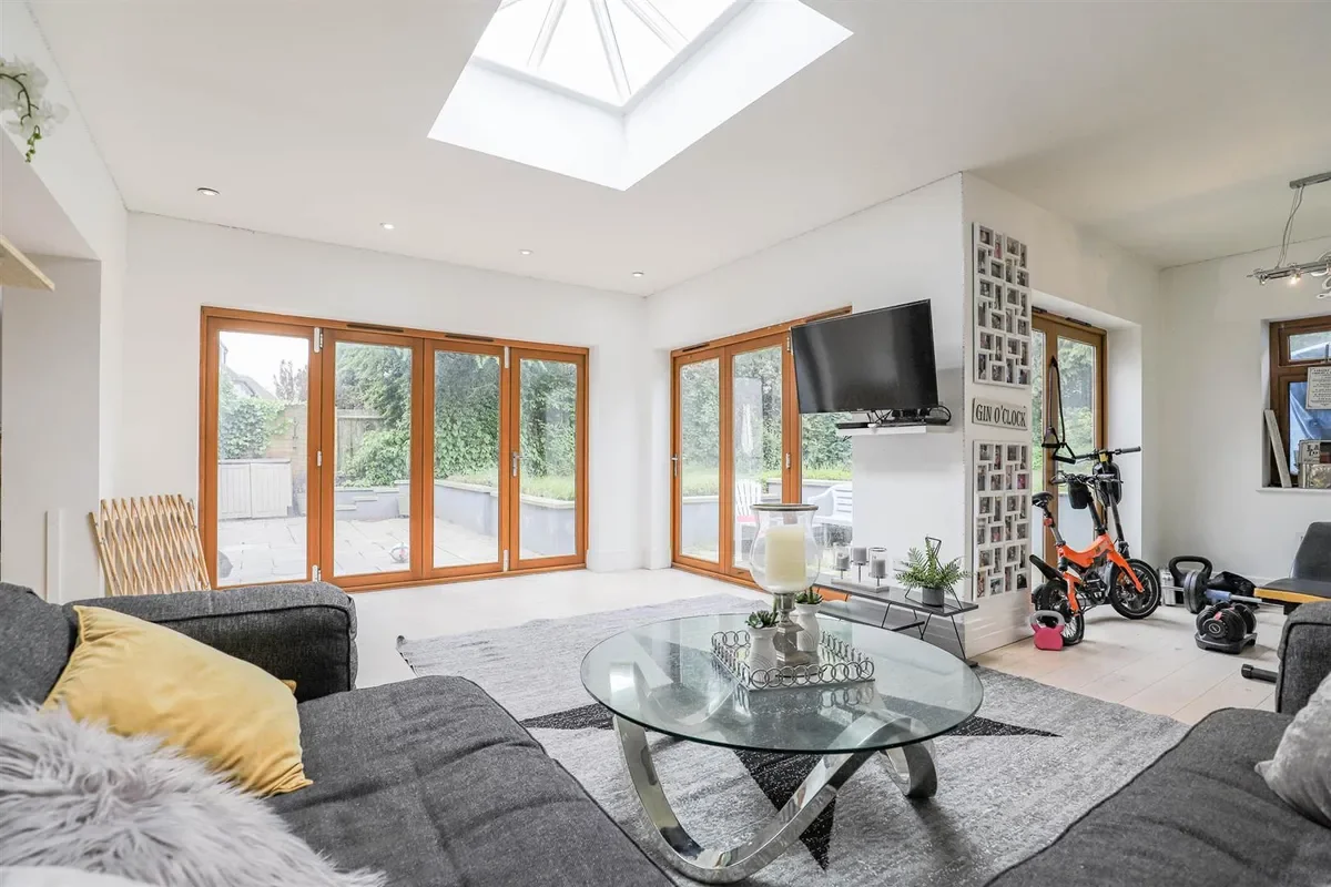 Living room with large glass sliding doors leading to a backyard, a gray sofa with yellow and gray pillows, a glass coffee table, a wall-mounted TV, a small exercise bike, and natural light from a skylight.