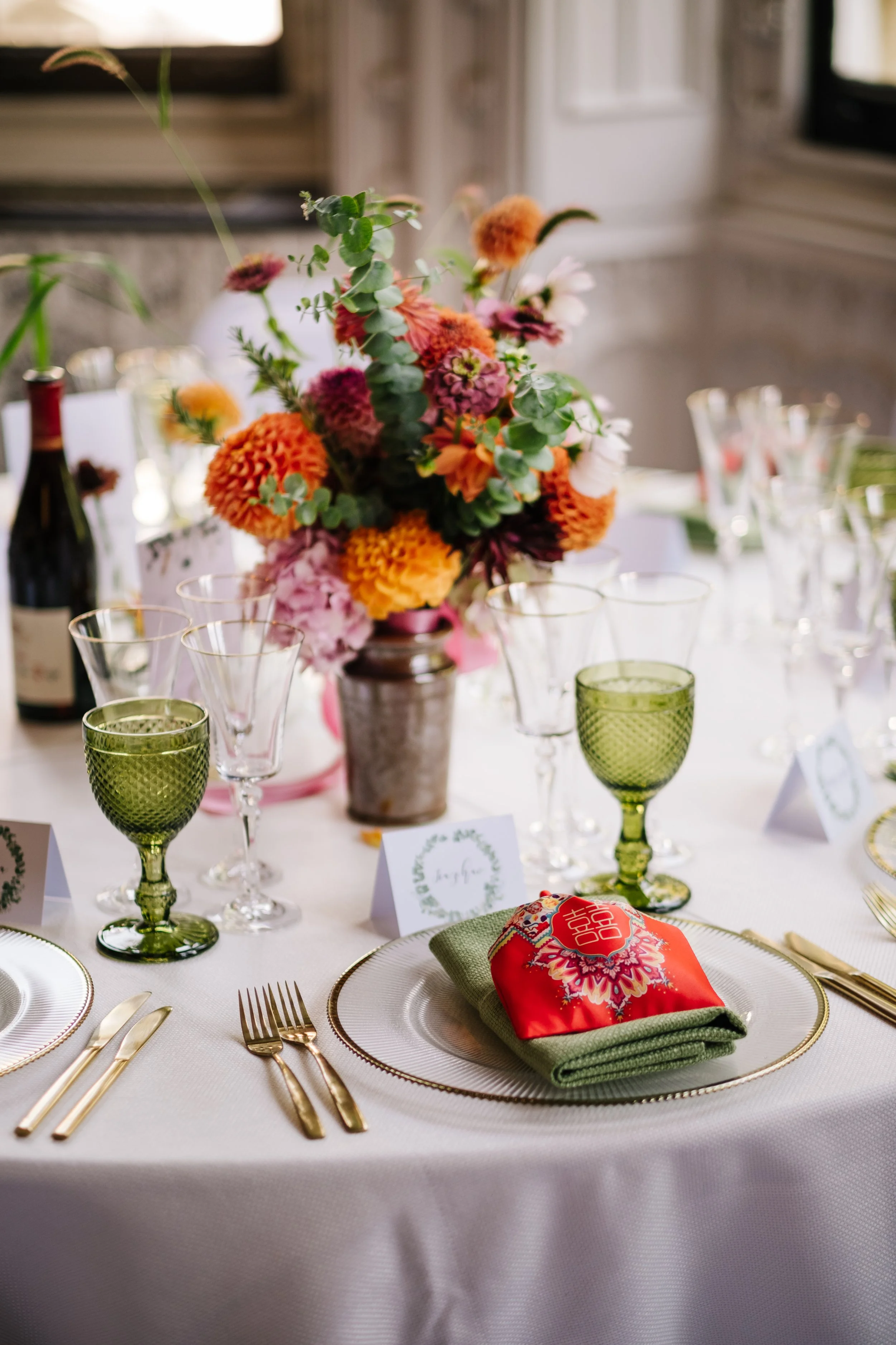 Wedding place setting with colourful floral centerpiece, green and clear glasses, gold cutlery, a white tablecloth.