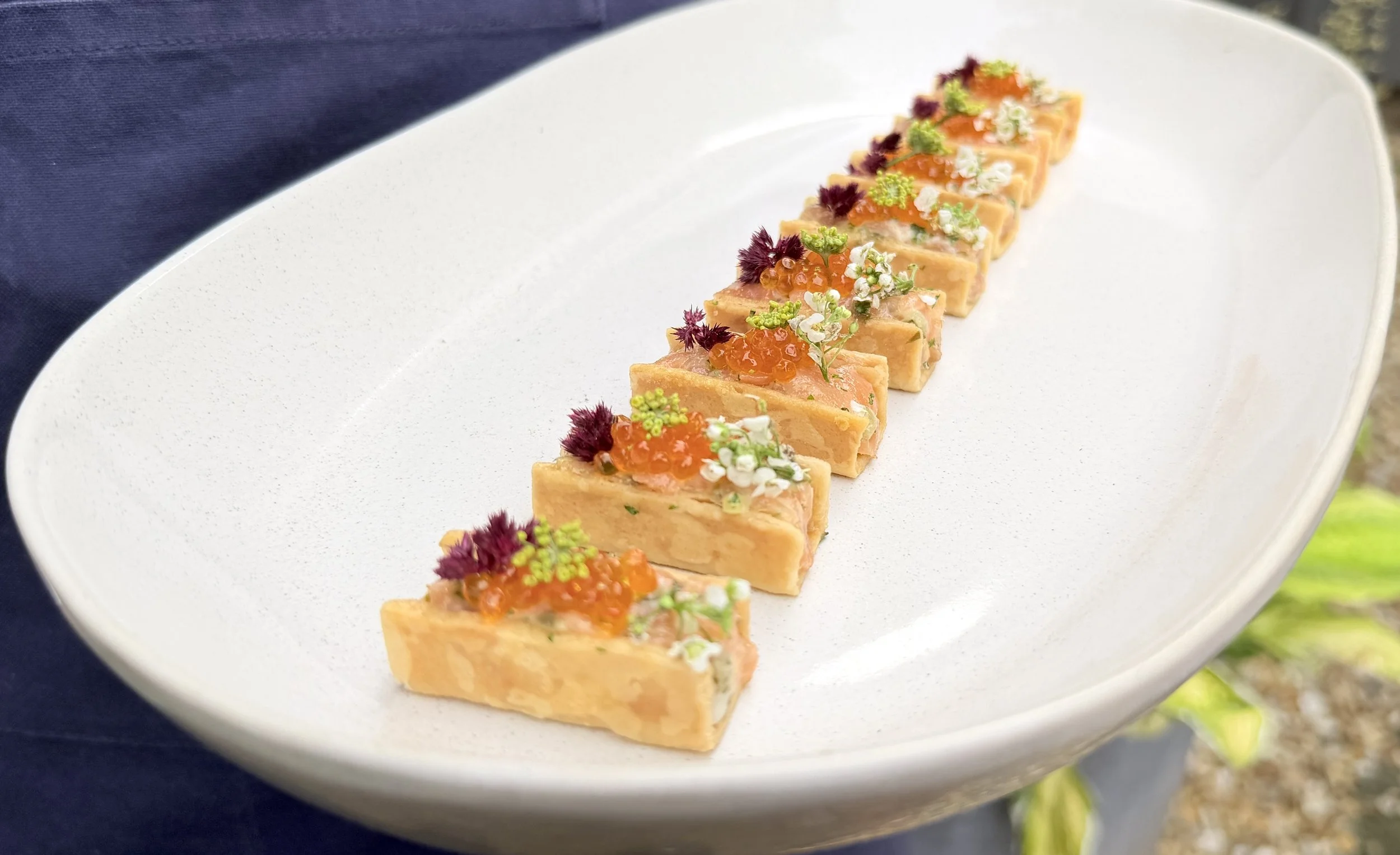 Sliced terrine topped with orange fish roe, small white and green flowers, and purple garnishes, arranged in a line on a white oval plate.