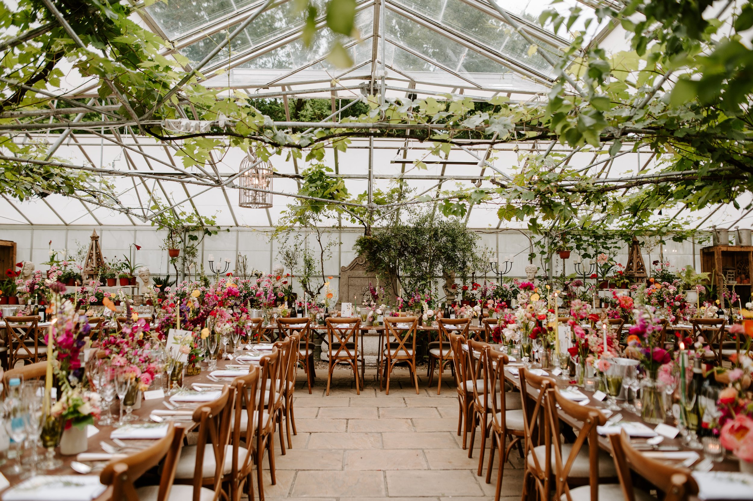 Indoor garden with a glass ceiling, decorated with numerous pink and white flowers, surrounded by wooden chairs and tables set for a celebration or event.