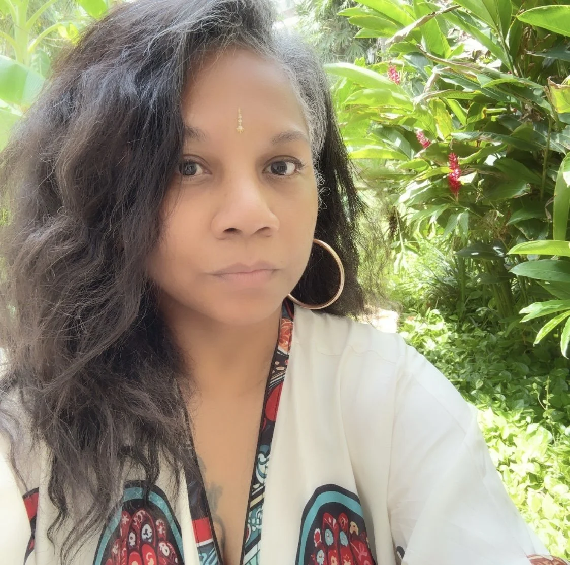 A woman with long, wavy black hair and large hoop earrings in a lush, green outdoor setting surrounded by tropical plants and pink flowers.