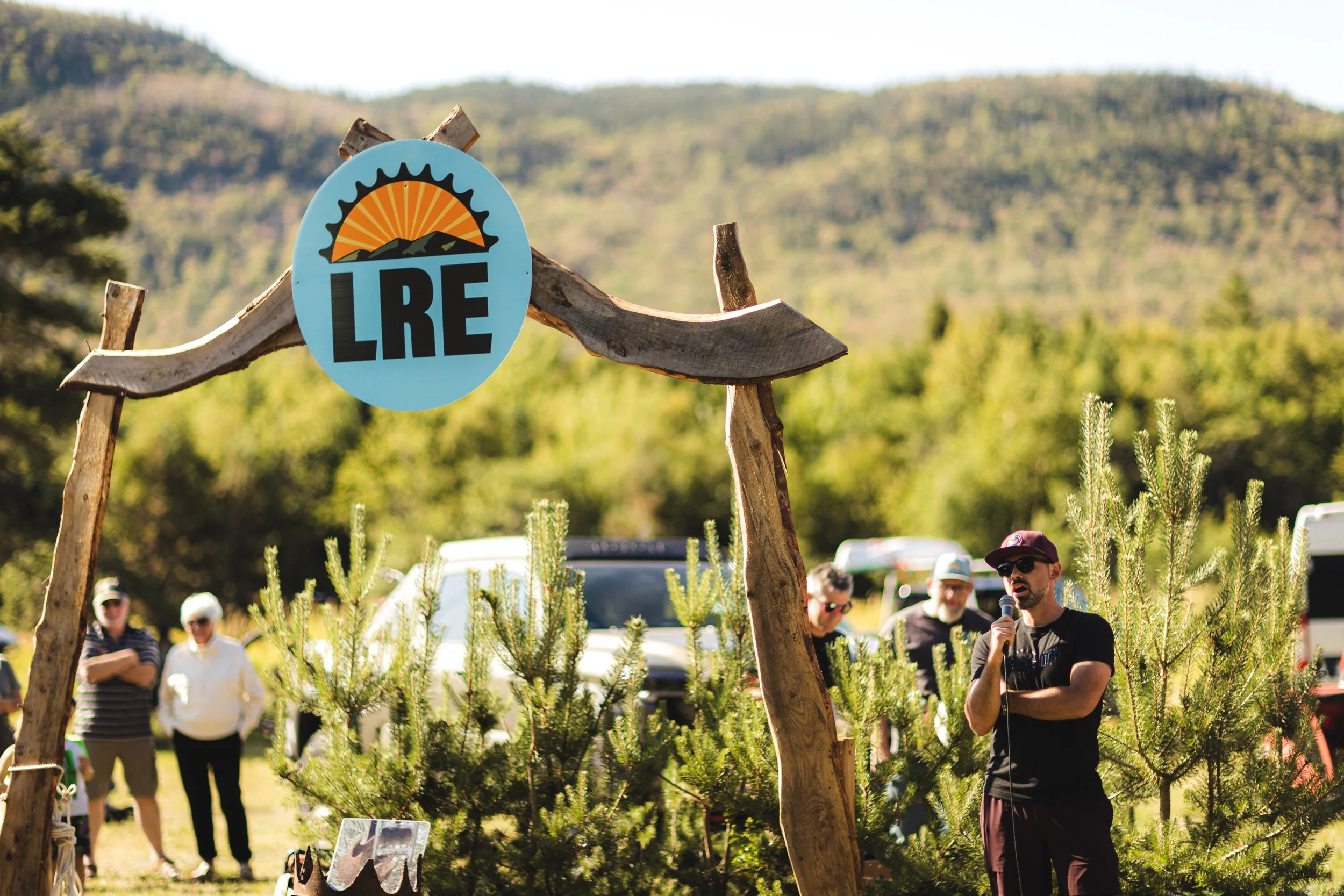 A man speaking into a microphone outdoors at a logging event, with a sign that reads "LRE" and a scenic background of mountains and trees.