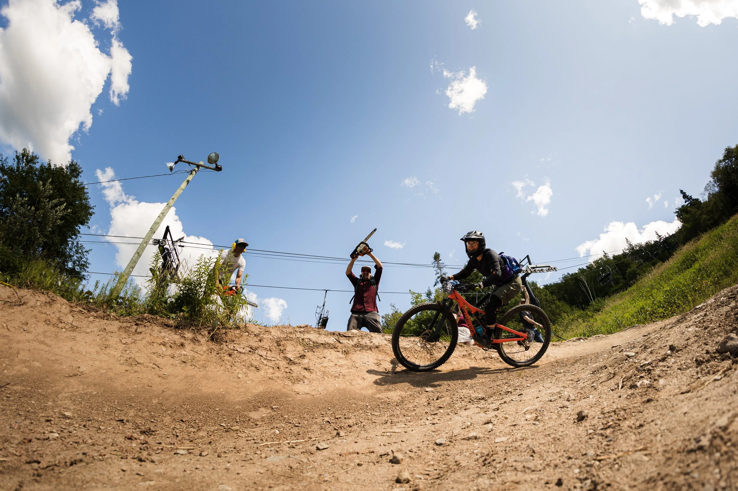 A mountain biker on a dirt trail during daytime, with two people standing on the side holding a sword and raising their hands. The sky is partly cloudy with a few trees and a ski lift in the background.