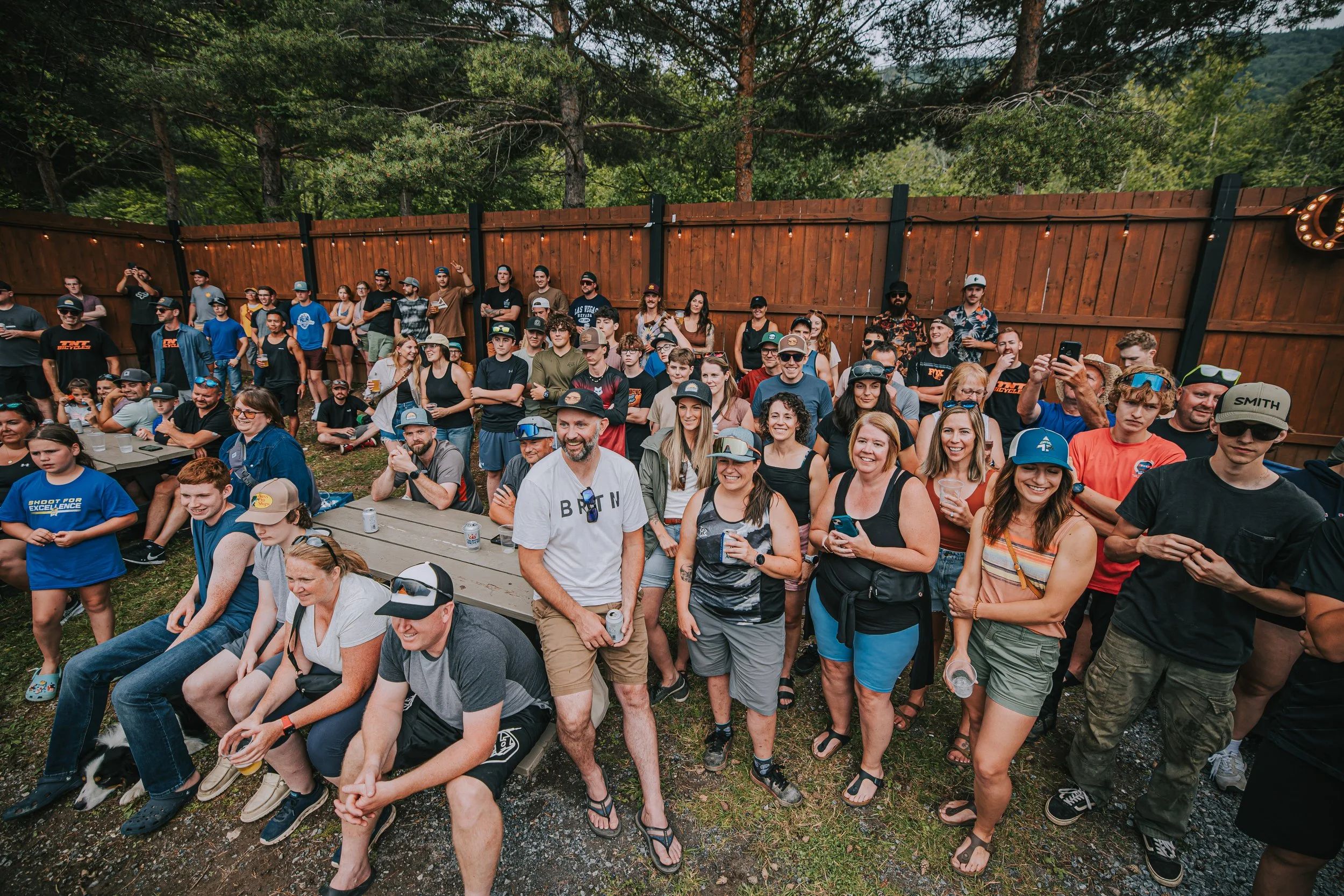 A group of people gathered outdoors in a fenced backyard, some sitting and some standing, enjoying a social event with drinks, surrounded by trees and string lights.