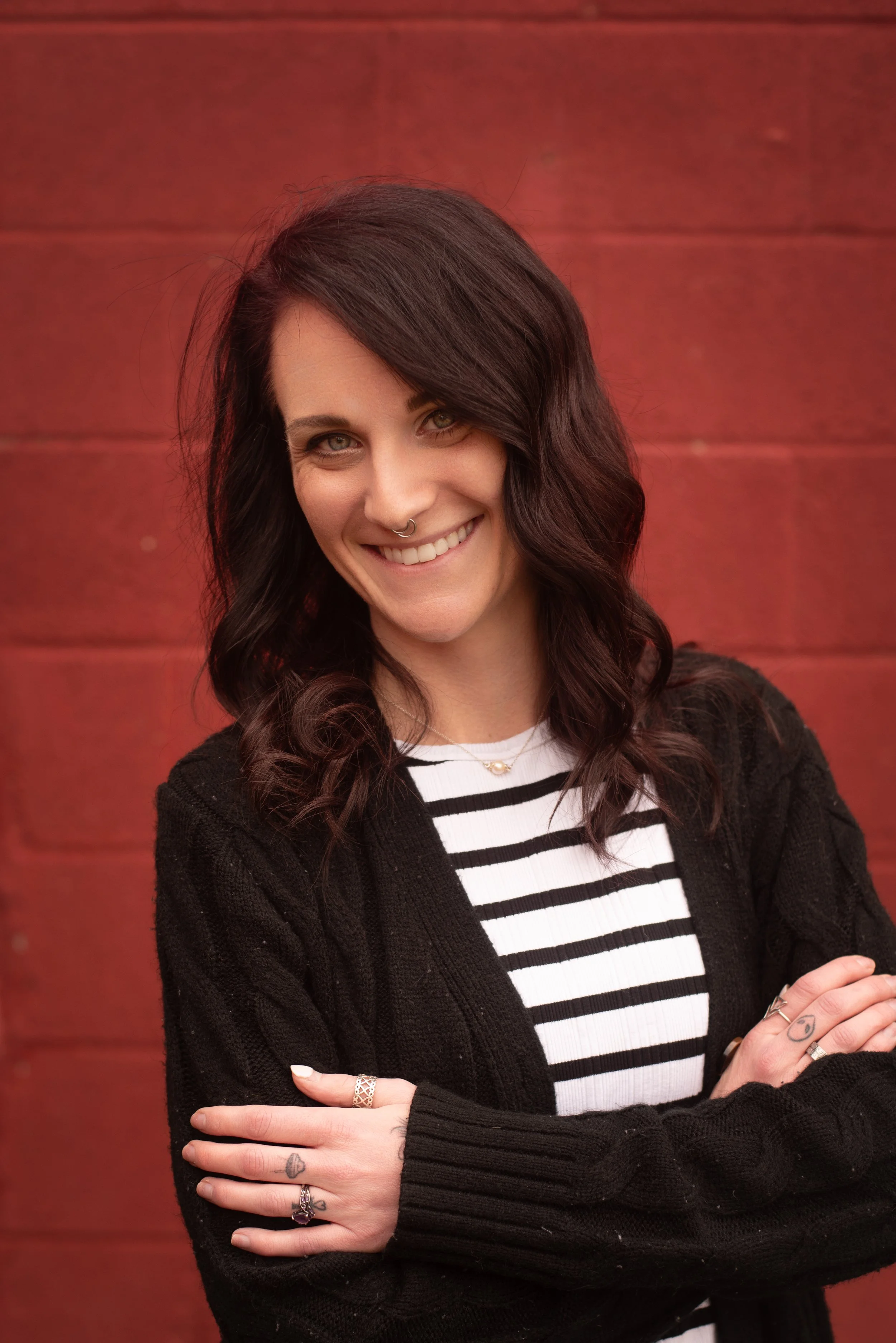 Woman with long dark hair and tattoos on her arms, crouching in a grassy field with yellow and orange flowers, smiling at the camera during sunset.