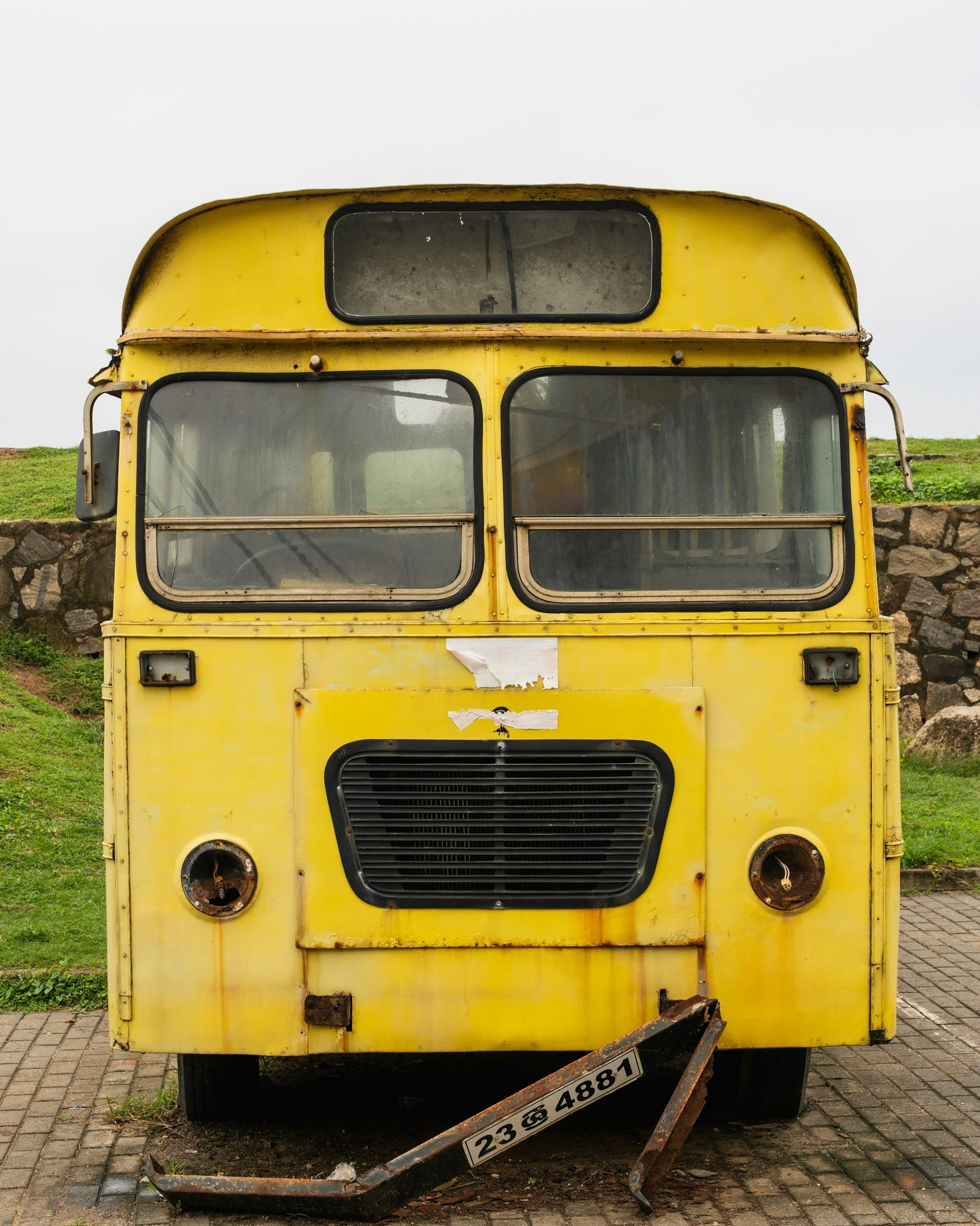 Front view of an old yellow bus with rust and damage, missing its headlights, parked on a paved sidewalk with grassy area and stone wall behind it.