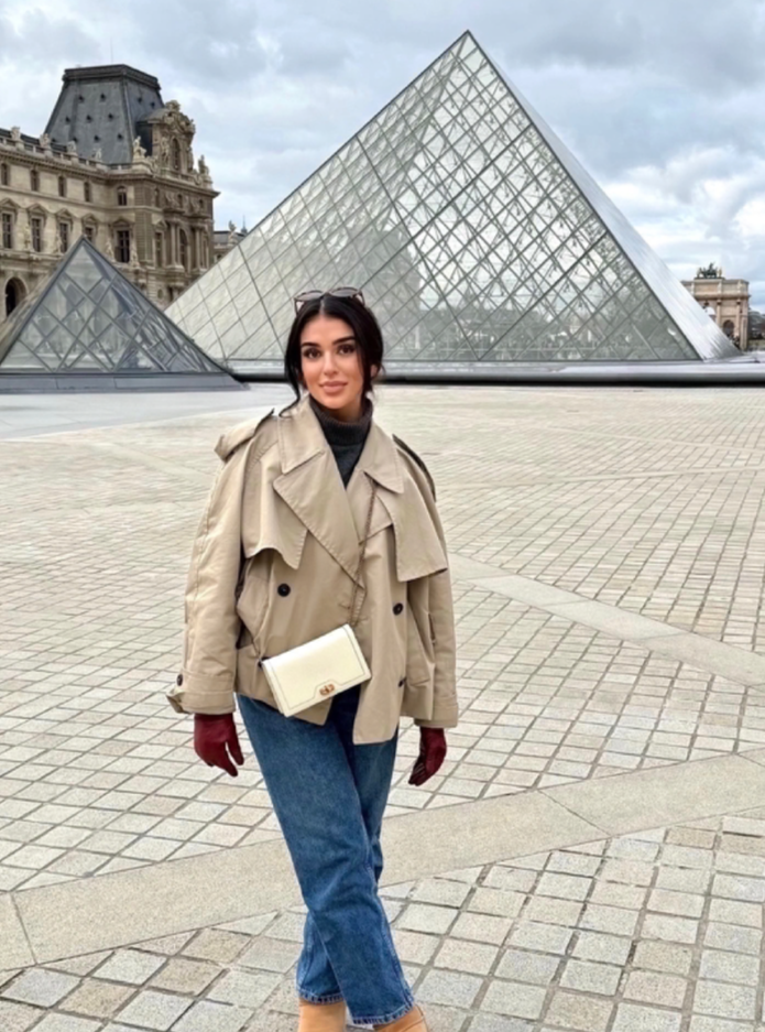 A woman standing in front of the glass pyramid entrance of the Louvre Museum in Paris, France. She is wearing a beige trench coat, red gloves, blue jeans, and tan shoes, with sunglasses resting on her head.