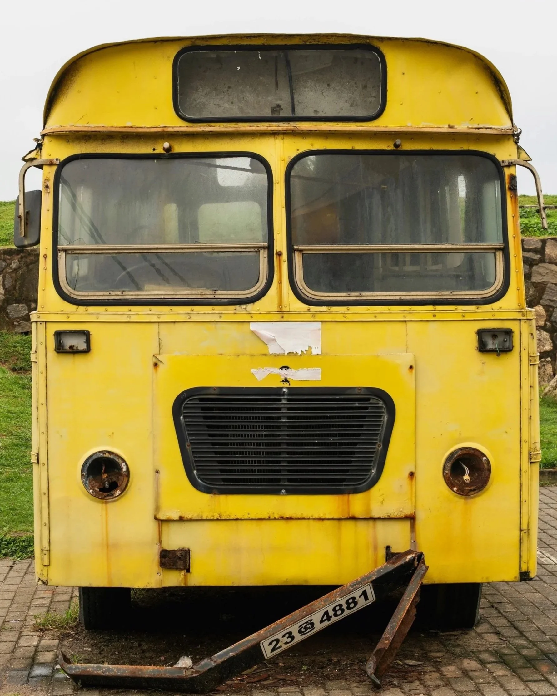 An old yellow bus with cracked and rusted parts, missing front headlights, and a broken bumper, parked on a brick pavement with grass and a stone wall in the background.