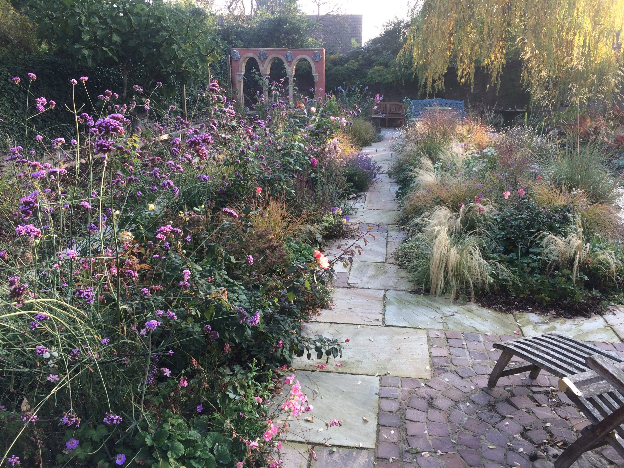 The view down the central sandstone pathway, framed either side by spectacular late summer blooms and soft textured grasses. A colonnade stands tall above the planting forming a focal point.
