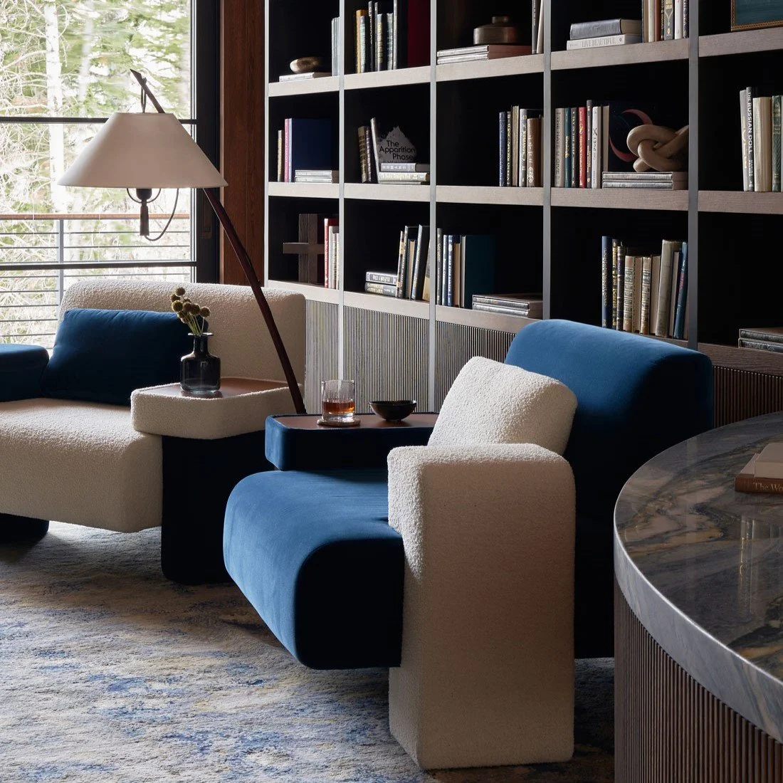 Sala de estar con sillones blancos y azules, lámpara de pie, librero con libros y decoración, ventana con vista a árboles.